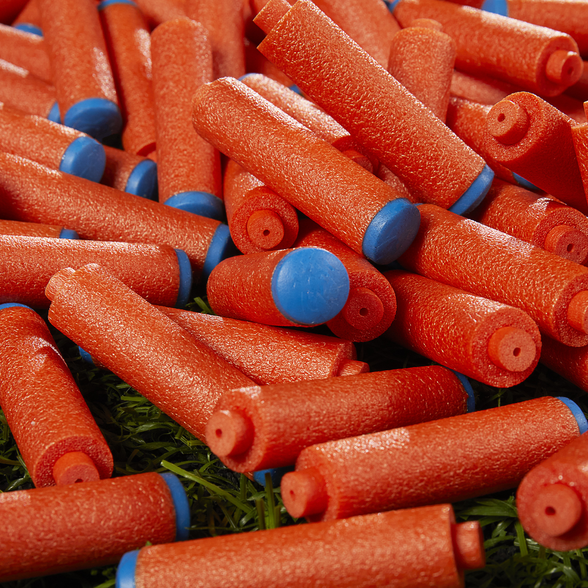 Primer plano de numerosas fideos de piscina de espuma roja con extremos azules, dispersos sobre césped artificial verde.