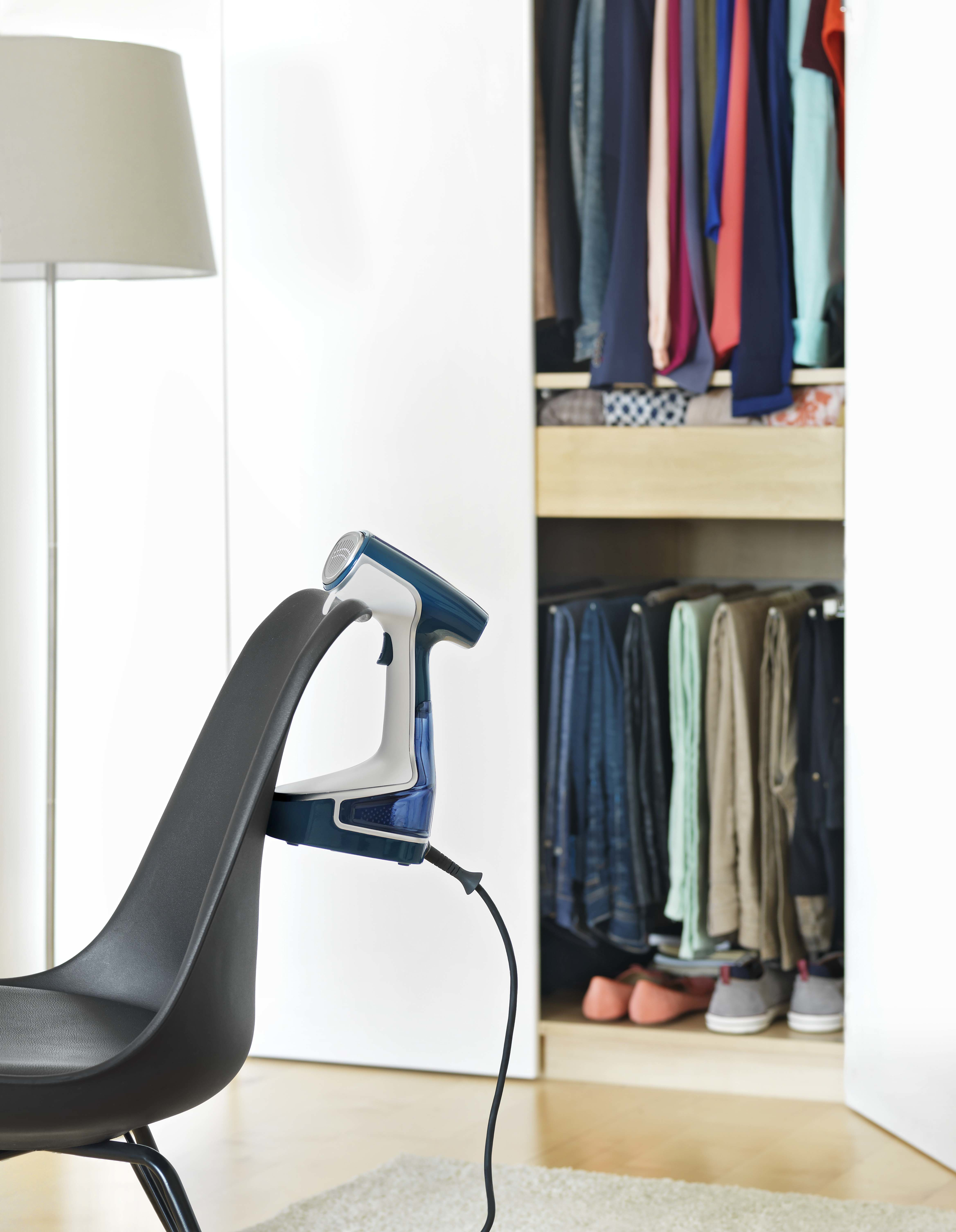 Un vaporizador de ropa azul y blanco está sobre una silla negra. La ropa cuelga en un armario abierto. Una lámpara de pie está a la izquierda.