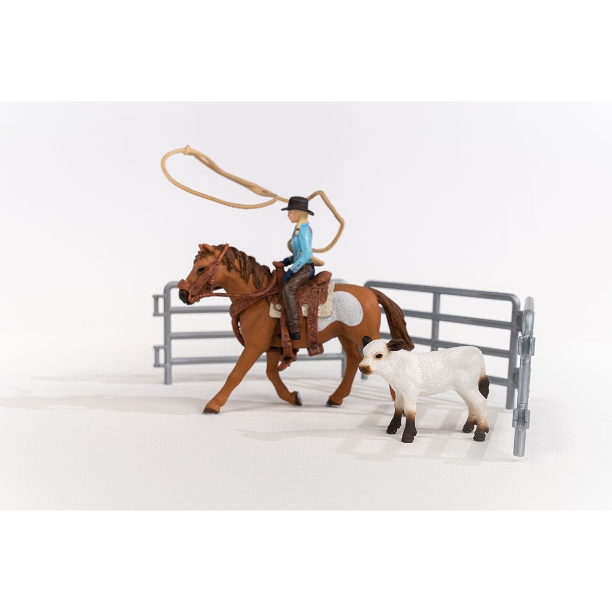 A toy scene shows a rider on a horse roping a calf near a gray fence. The horse is brown, and the calf is white with dark spots.