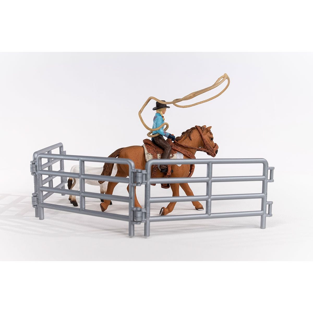 A toy cowboy on a brown horse with a rope, inside a metal fence, with a white horse in the background.