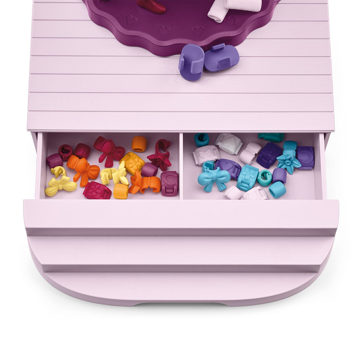 A pink toy box with open drawer. Inside are small colorful accessories like bows, flowers, and other shapes. Purple lid with heart shapes.
