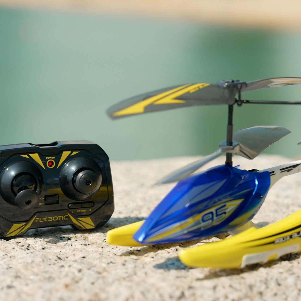 Ein Spielzeughelikopter mit blauen und gelben Details neben einer schwarzen Fernbedienung, vor verschwommenem Wasserhintergrund.