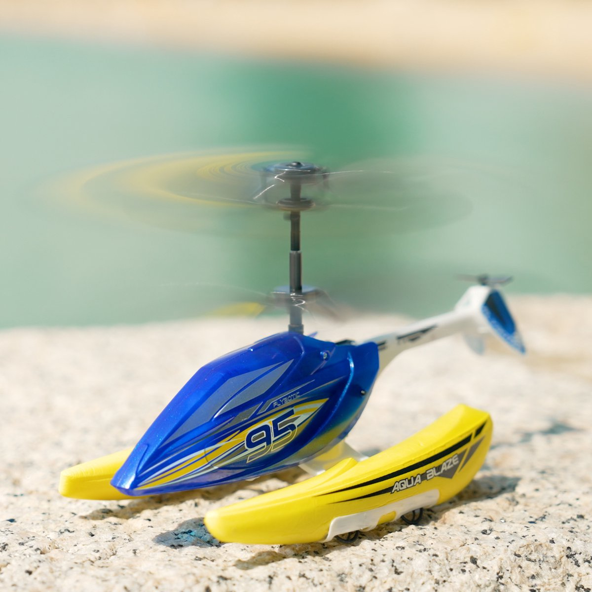 Ein blau-gelber Spielzeughelikopter mit sich drehenden Rotorblättern steht auf einer Steinoberfläche in der Nähe von Wasser.