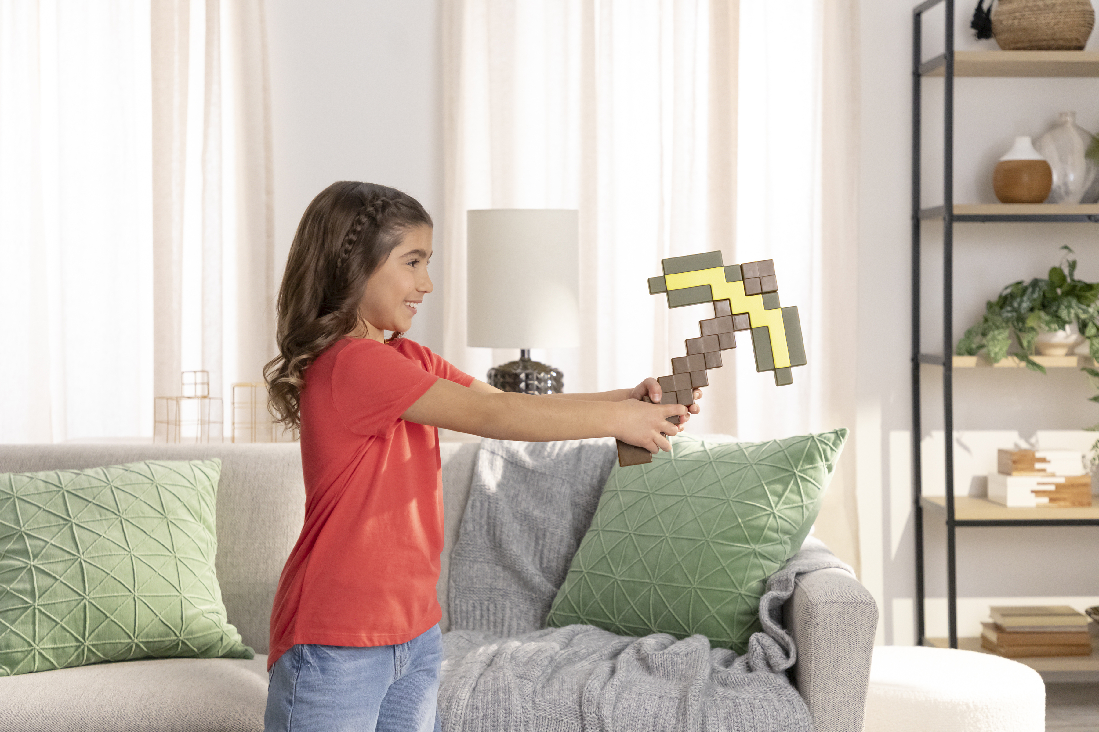 Dziewczyna trzyma kilof Minecraft. Ma na sobie czerwoną koszulę. Kanapa i półki w tle.