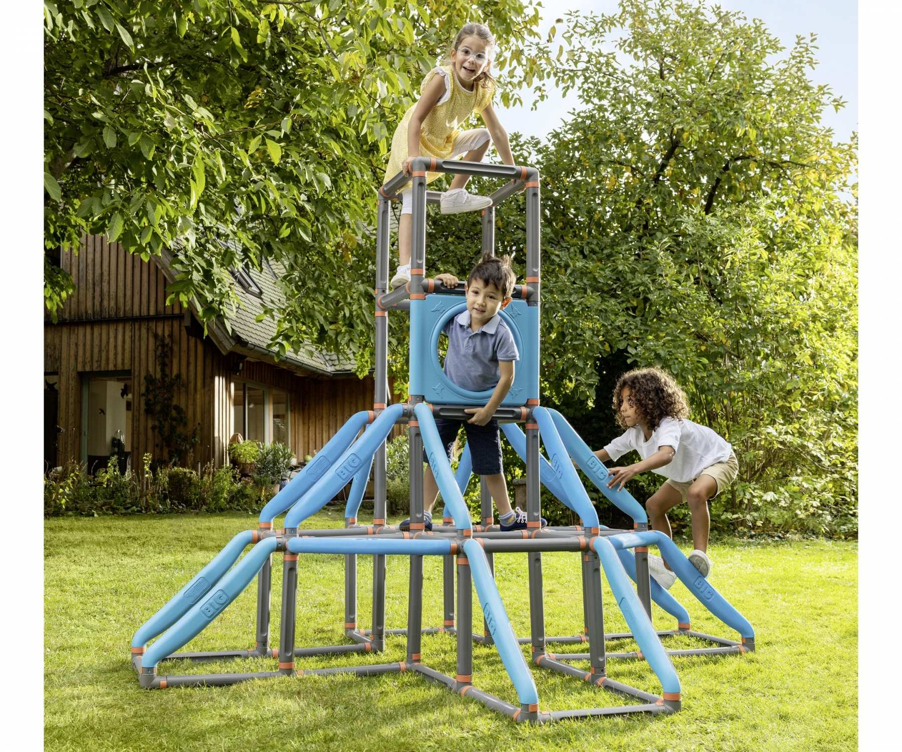 Drei Kinder spielen auf einem blau-grauen Spielgerüst in einem grasbewachsenen Garten. Bäume und ein Haus sind im Hintergrund.