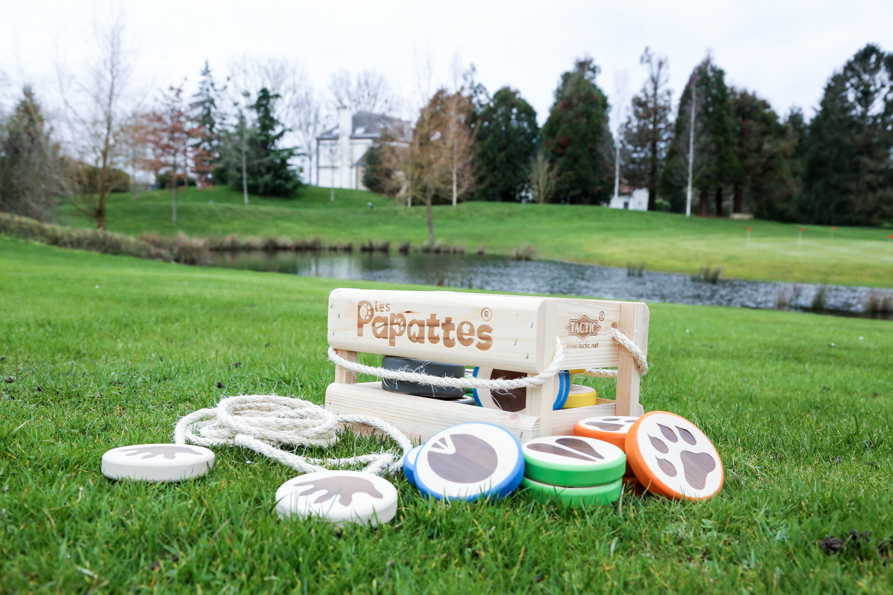 Juego de madera 'les Papattes' sobre hierba, discos con huellas, caja de madera, cuerda, estanque, árboles, casa detrás.