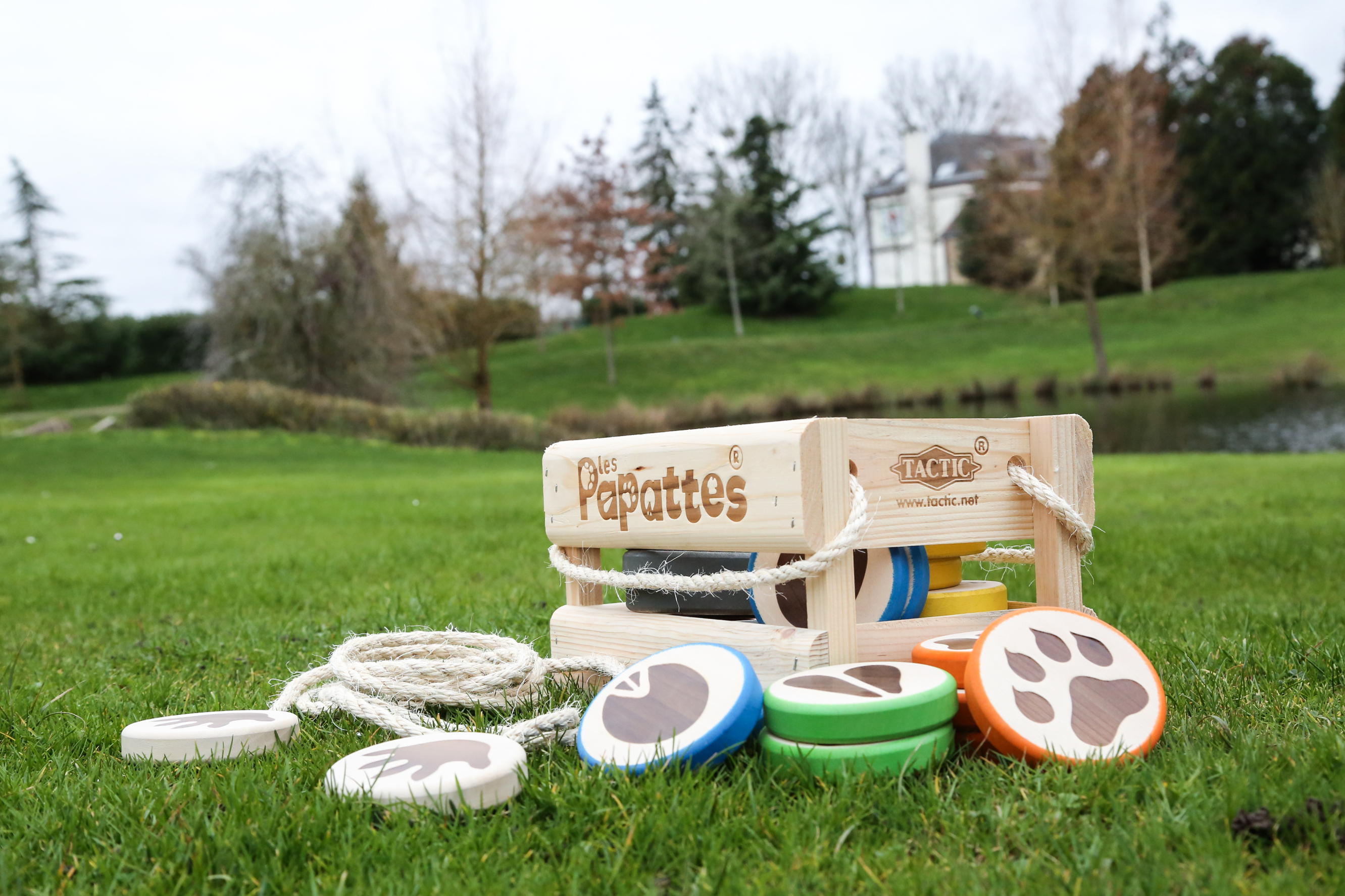 Juego de madera 'Les Papattes' con discos coloridos y cuerda sobre hierba. Árboles y un lago al fondo.