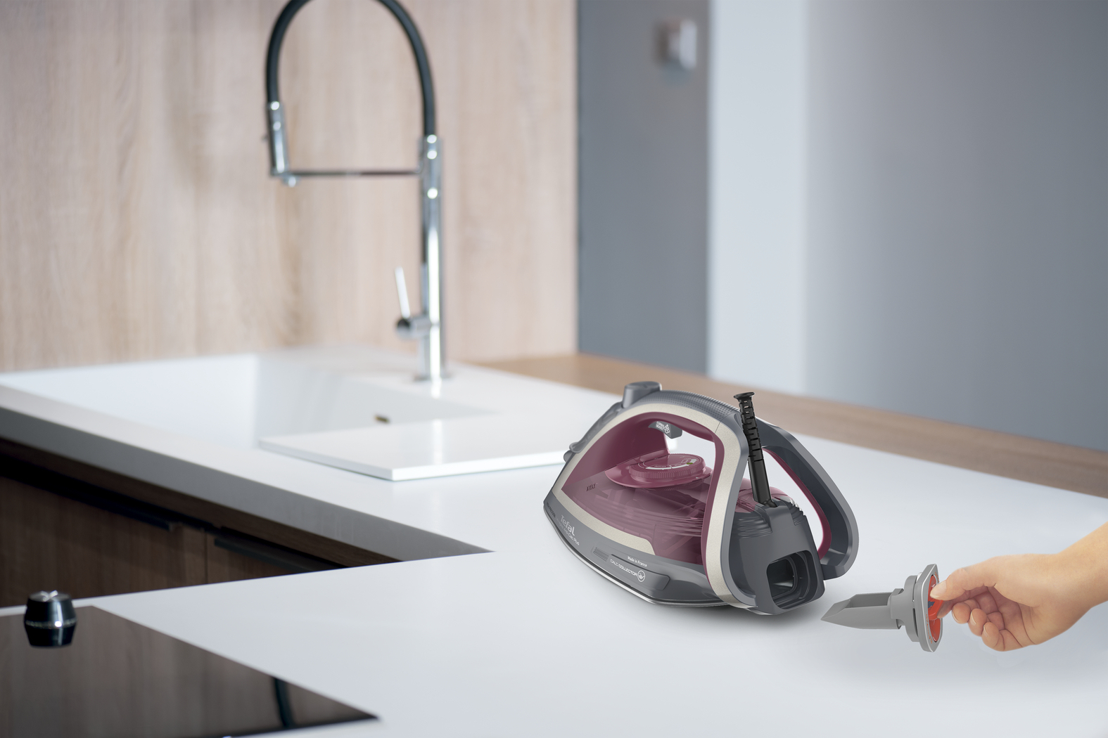 Żelazko na blacie. Ręka trzyma filtr. Kran i zlew z tyłu. Widoczne kolory szary, biały i bordowy.