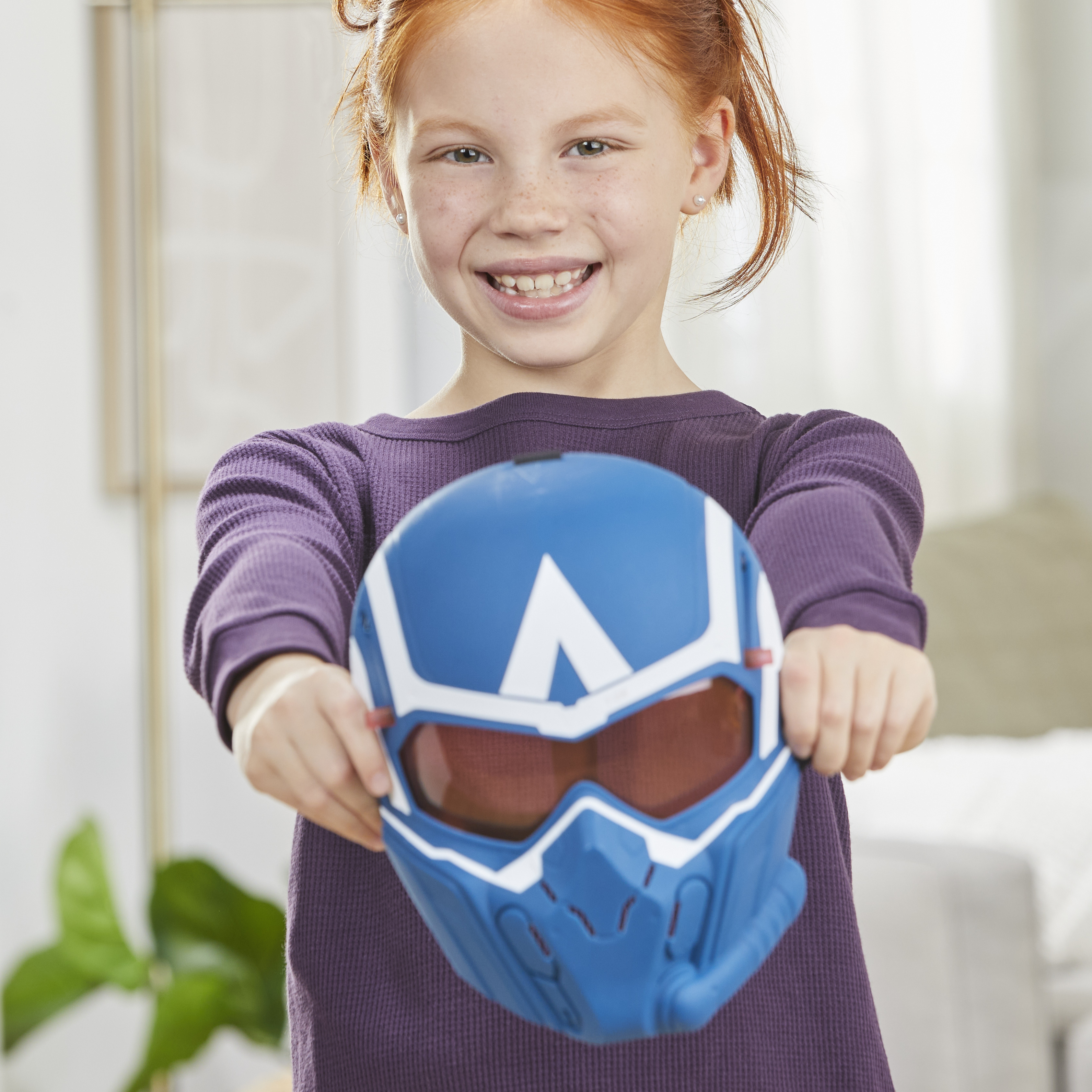 Una niña pelirroja sostiene una máscara azul y blanca. Ella sonríe. La máscara tiene lentes tintados rojos.