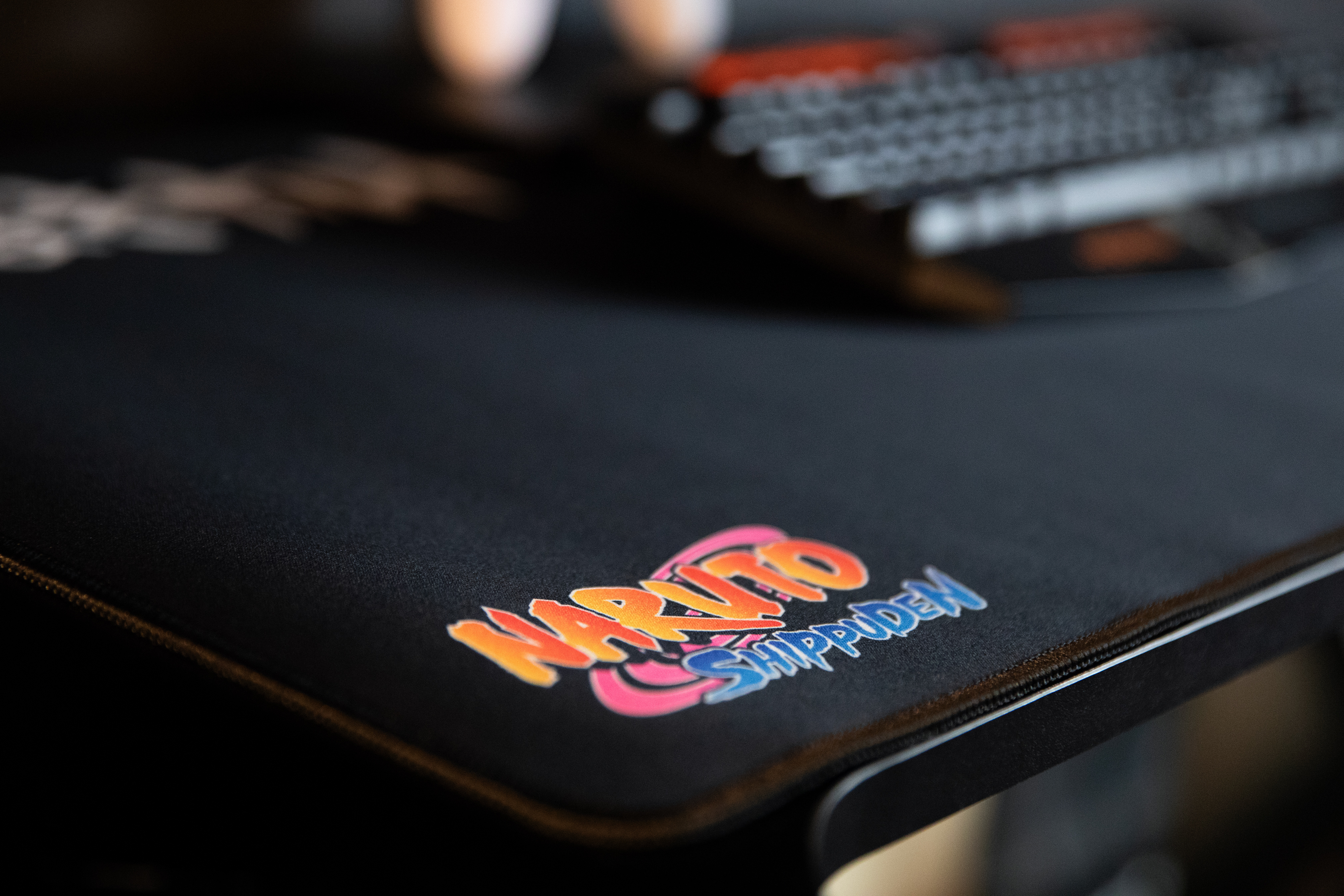 Zbliżenie czarnej podkładki pod mysz z logo Naruto Shippuden w żywych kolorach. Klawiatura jest lekko widoczna w tle.