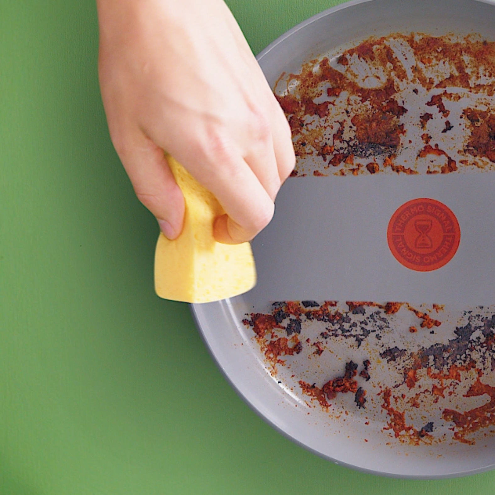 Ręka trzymająca żółtą gąbkę czyści brudną, szarą patelnię z czerwonym logo na zielonym tle.