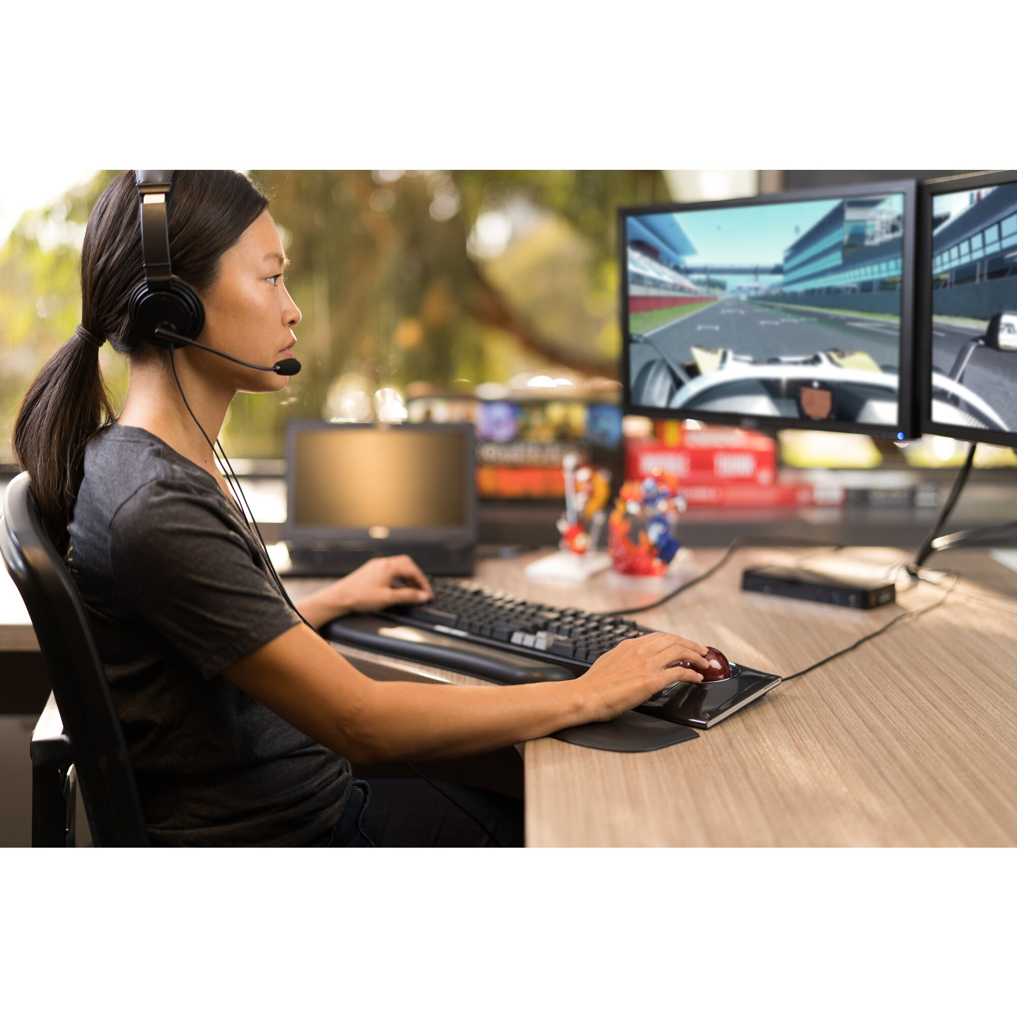 Una mujer con auriculares juega en una computadora. Dos monitores muestran un juego de carreras. Usa teclado y ratón.