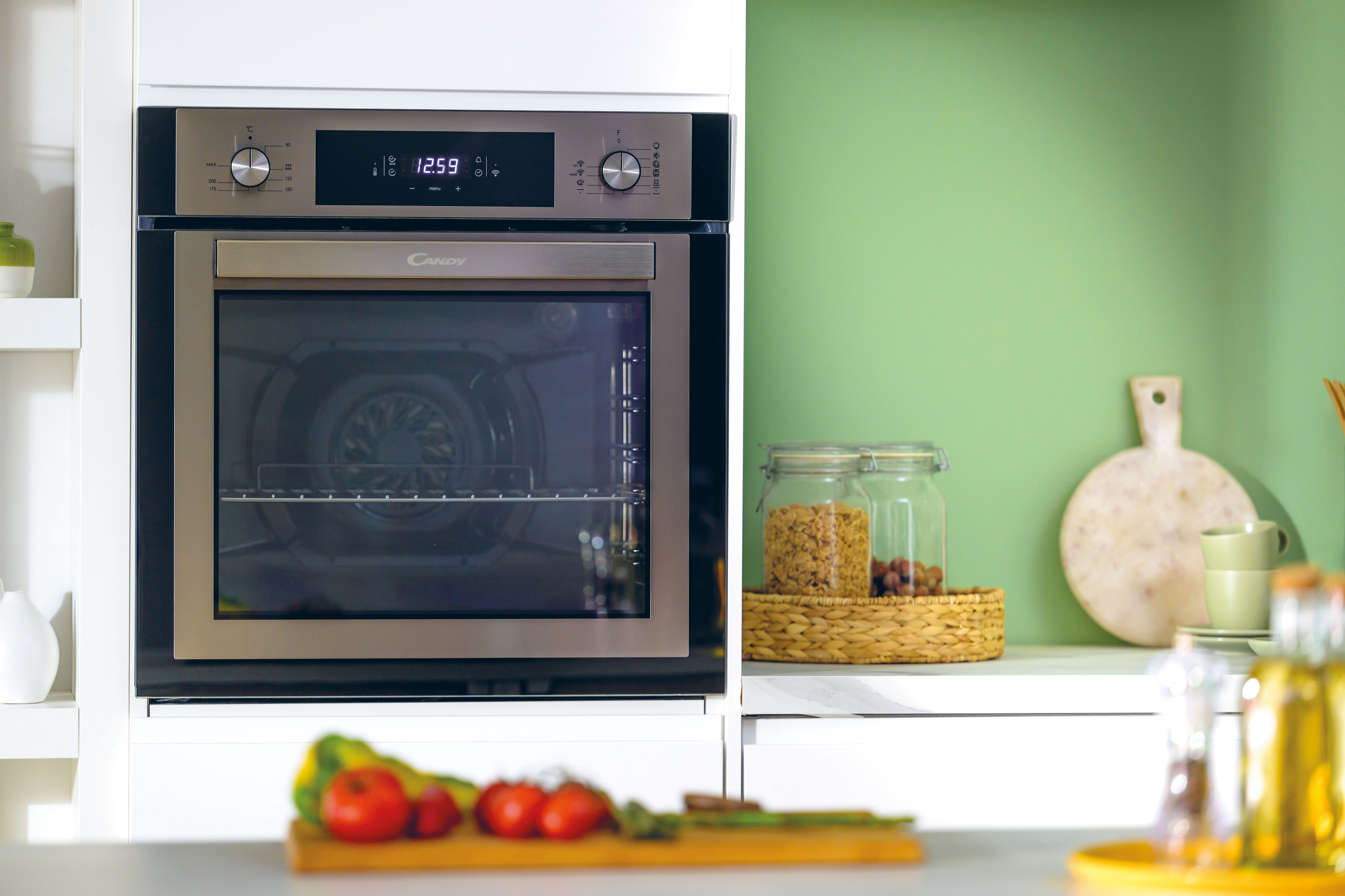 Una scena di cucina con forno, verdure e barattoli. Il forno è argentato, il muro è verde e il piano di lavoro è bianco.