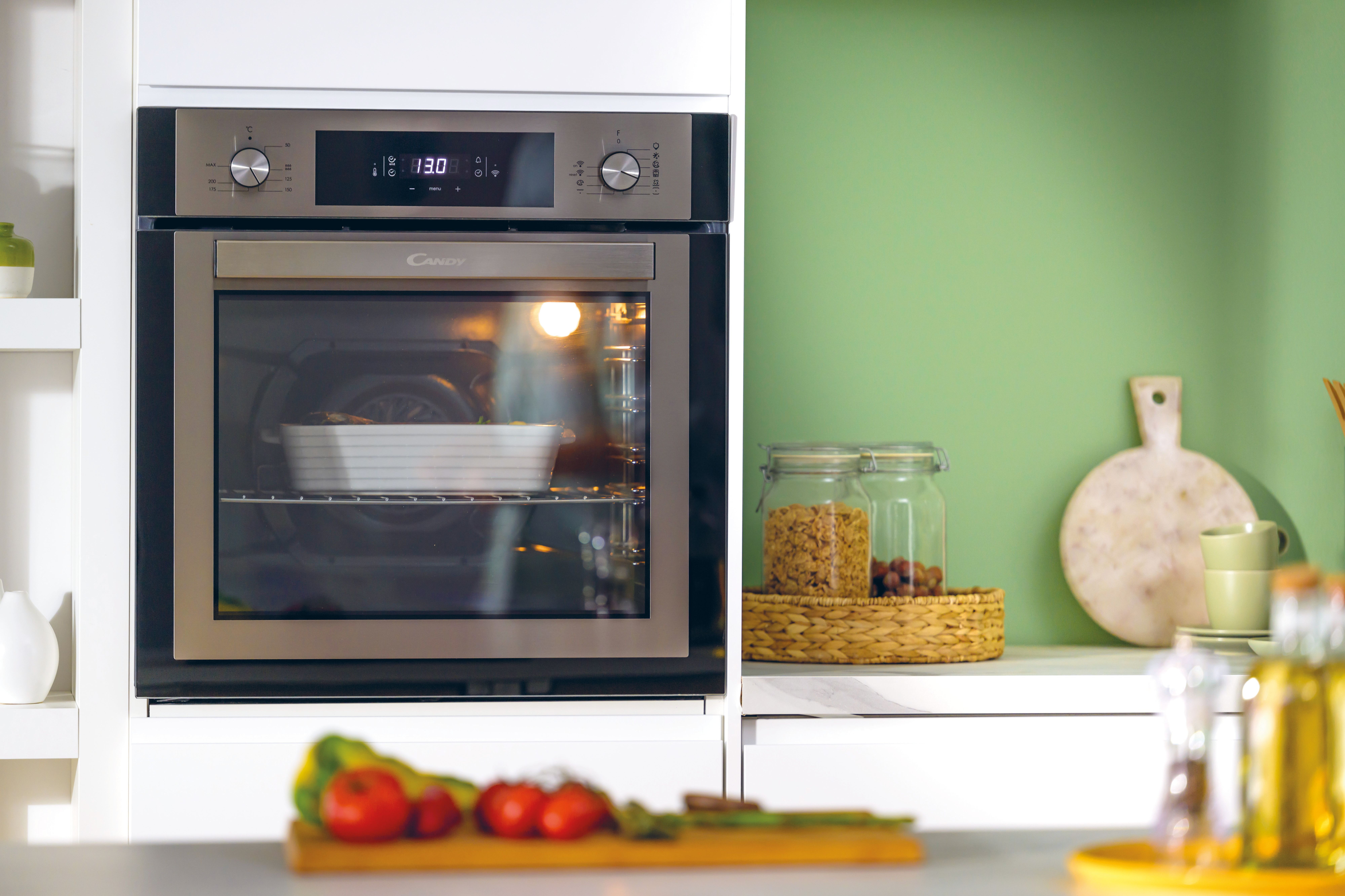 Un forno con un piatto all'interno, un piano cucina con cibo e barattoli, parete verde e armadi bianchi.
