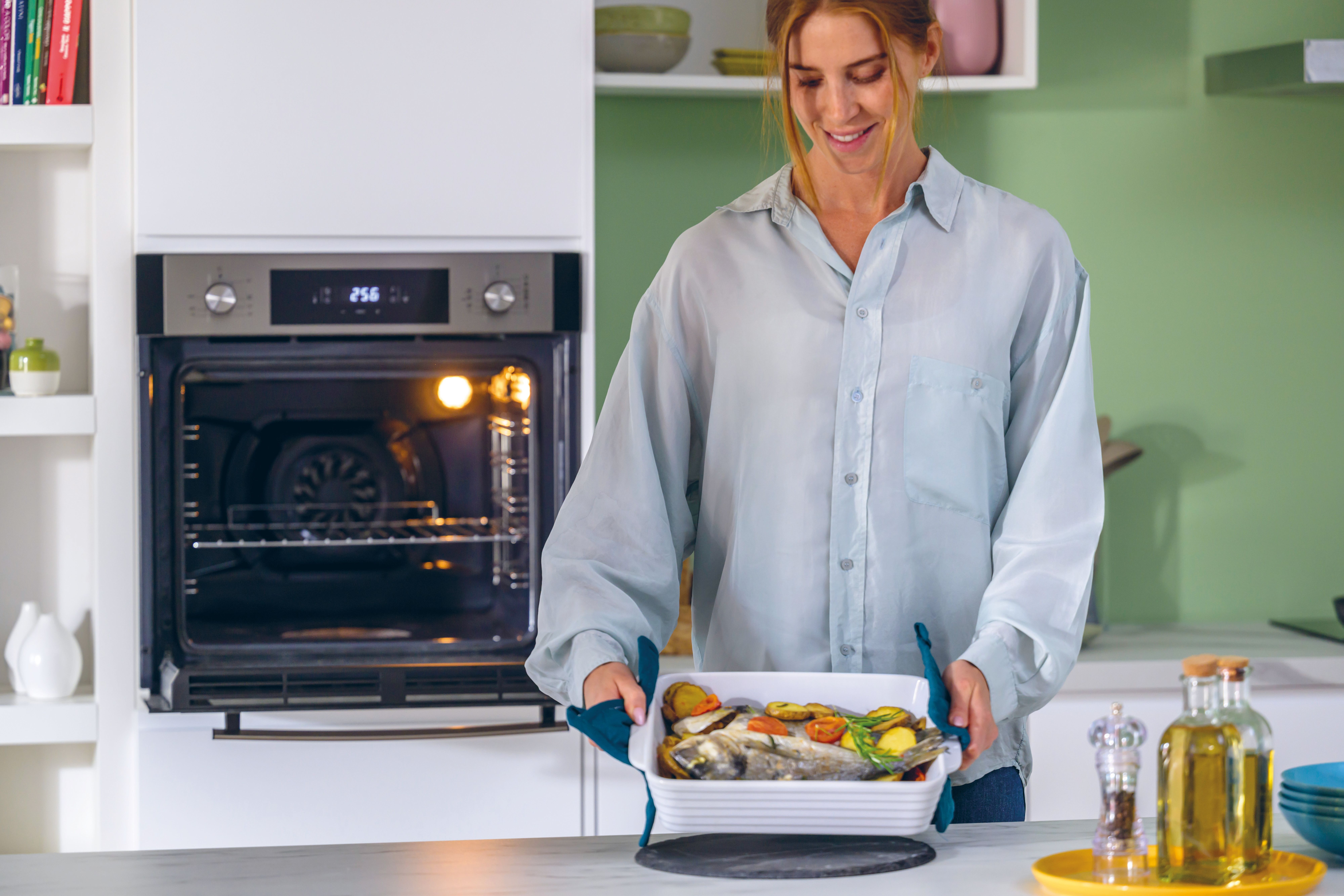 Donna che tiene un piatto di pesce al forno. Forno sullo sfondo. Ambiente cucina con olio d'oliva e ciotole.