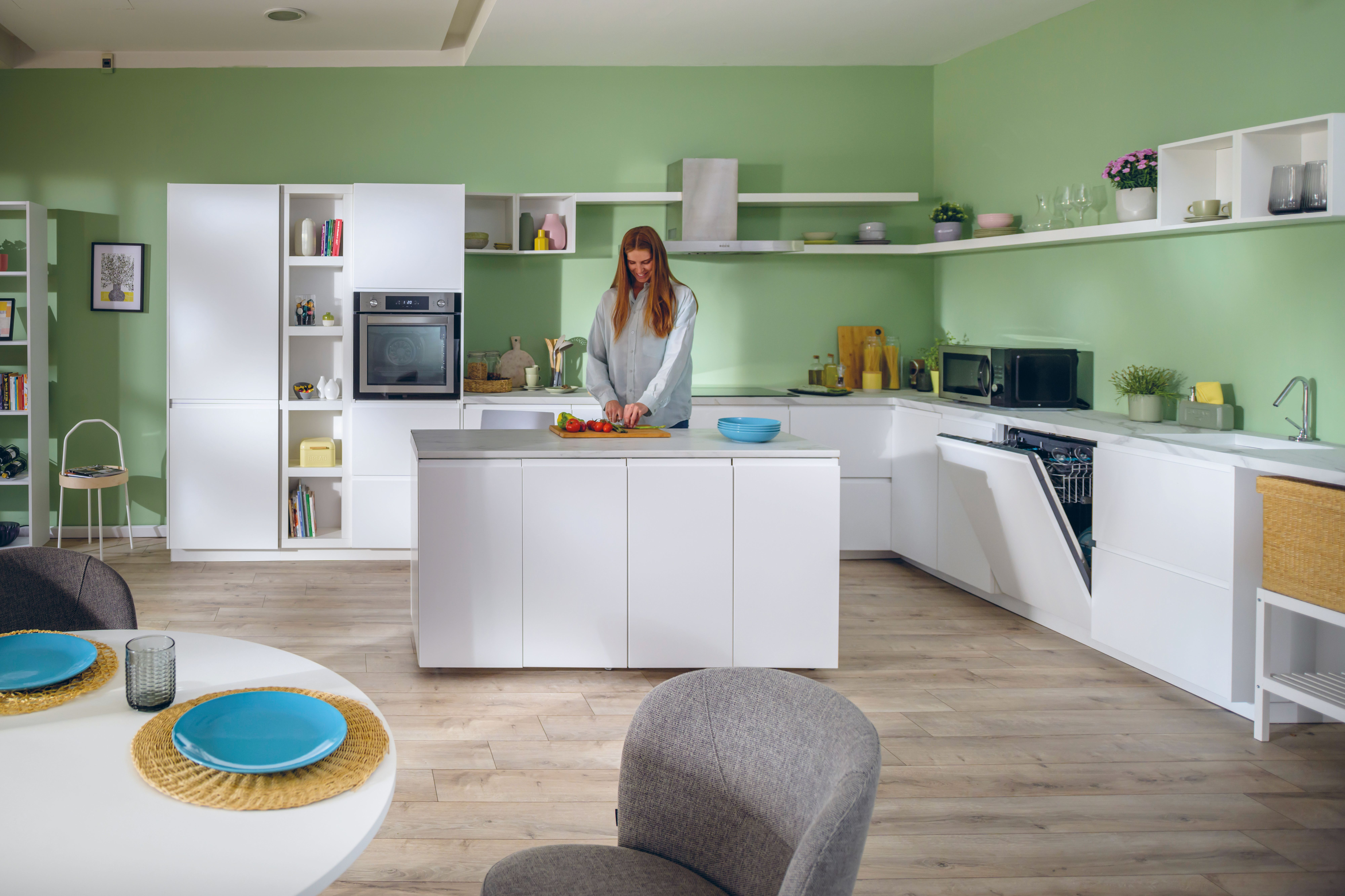 Una donna taglia le verdure in una cucina moderna. Armadi bianchi, pareti verdi e un tavolo rotondo sono visibili.