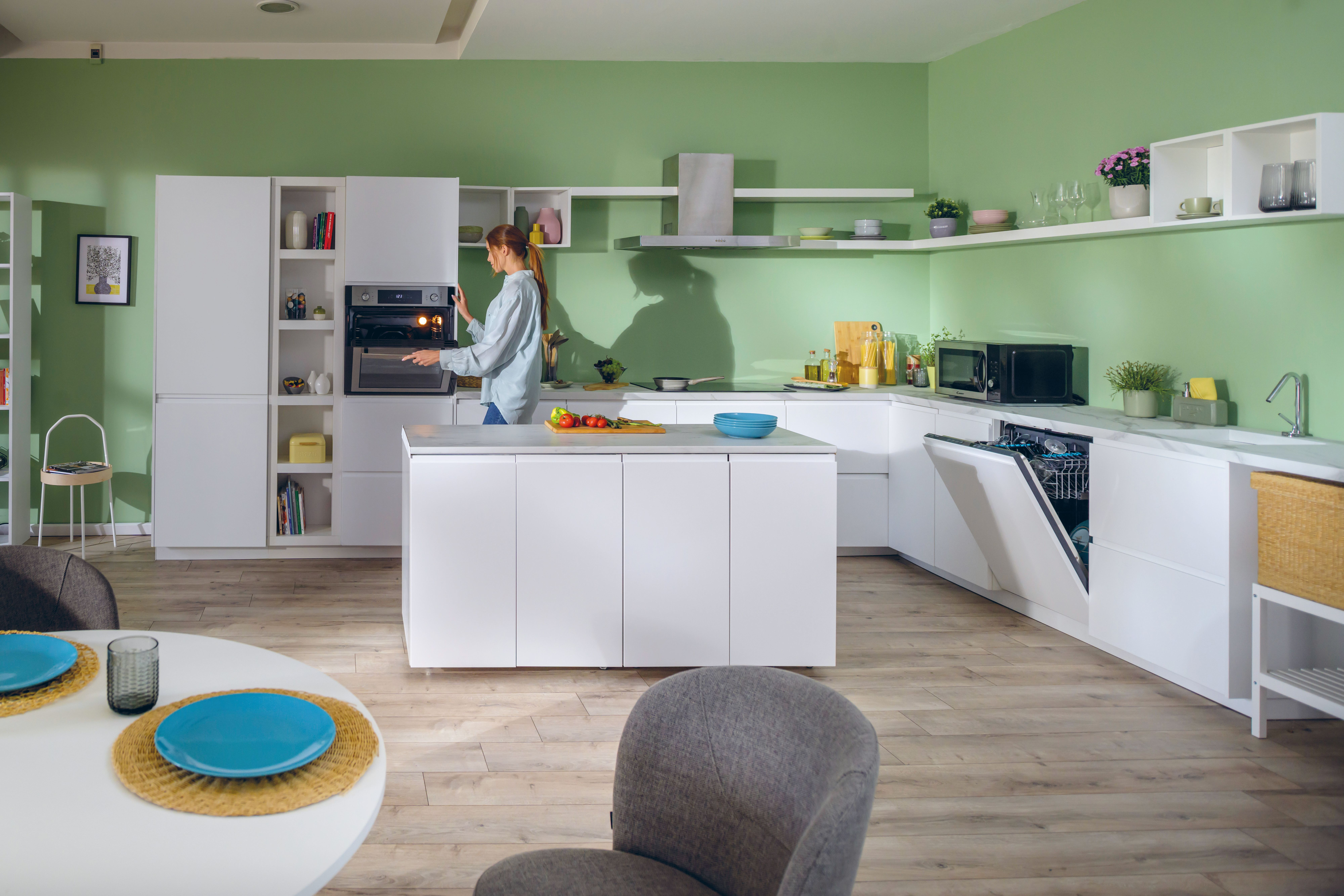 Una donna in una cucina moderna apre un forno. La cucina ha mobili bianchi, pareti verdi e un tavolo rotondo.
