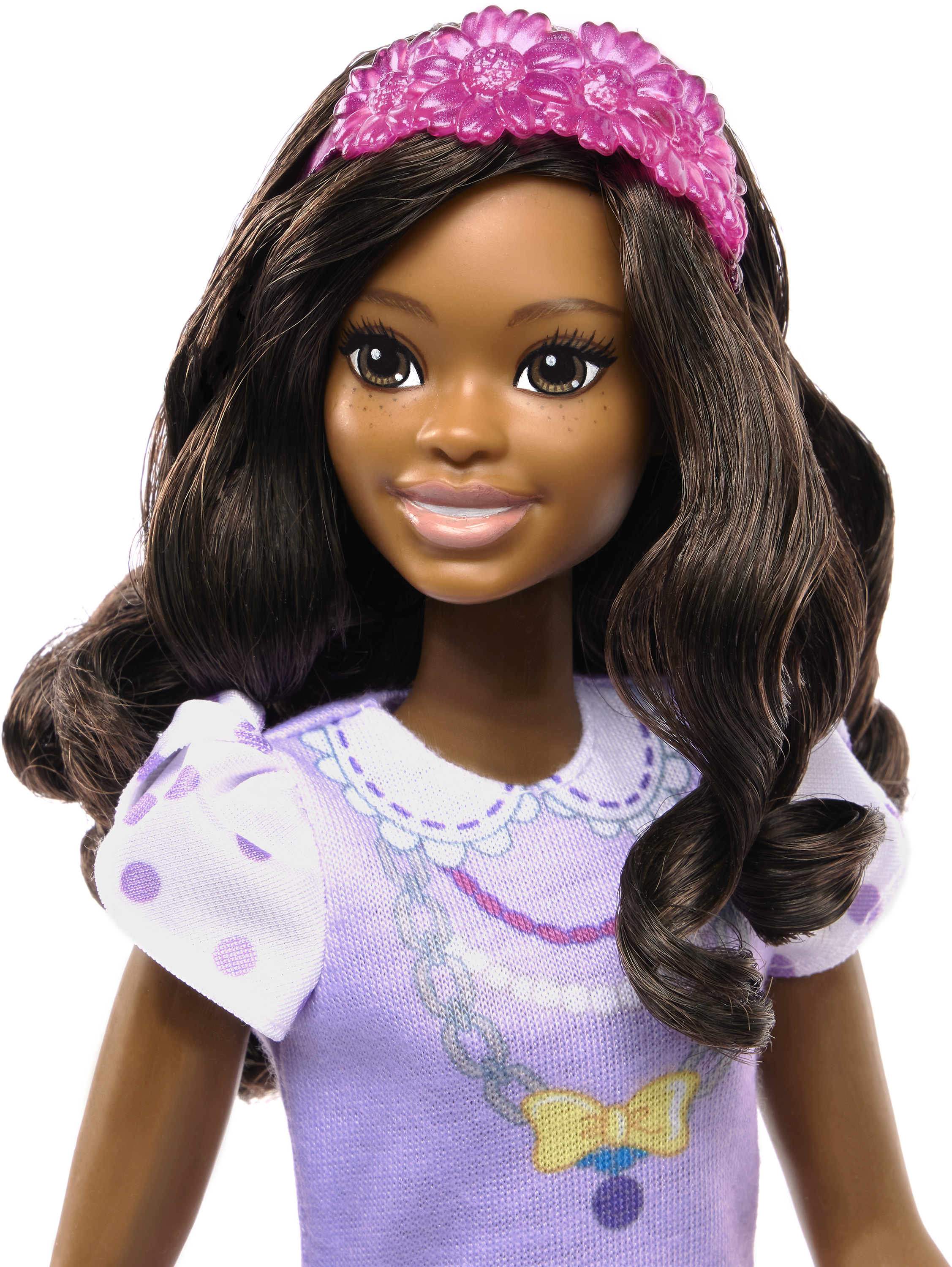Muñeca con cabello castaño y piel oscura, usando una diadema rosa y un vestido morado con lunares blancos.