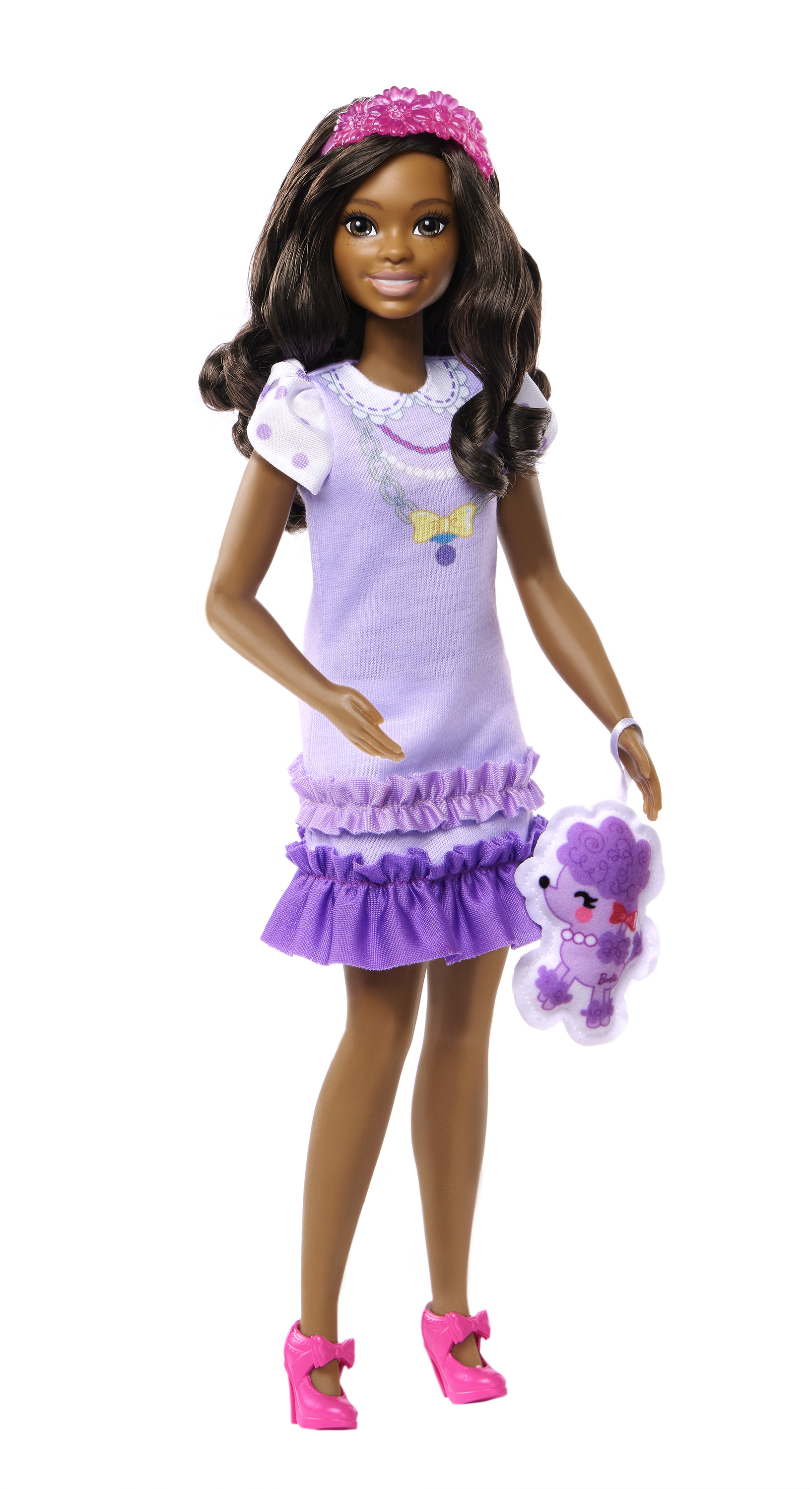 Una muñeca de piel oscura, con un vestido morado y zapatos rosas. Sostiene un caniche de juguete. Una corona de flores rosas en la cabeza.
