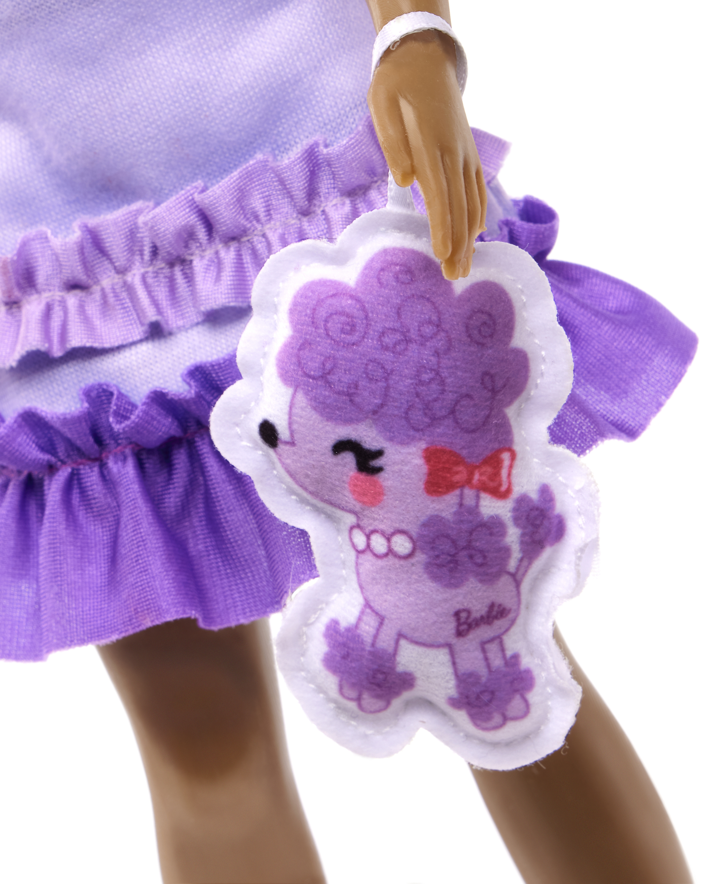 Una muñeca sostiene un bolso con forma de caniche. La muñeca usa un vestido morado y tiene piel oscura. El bolso es blanco y morado.