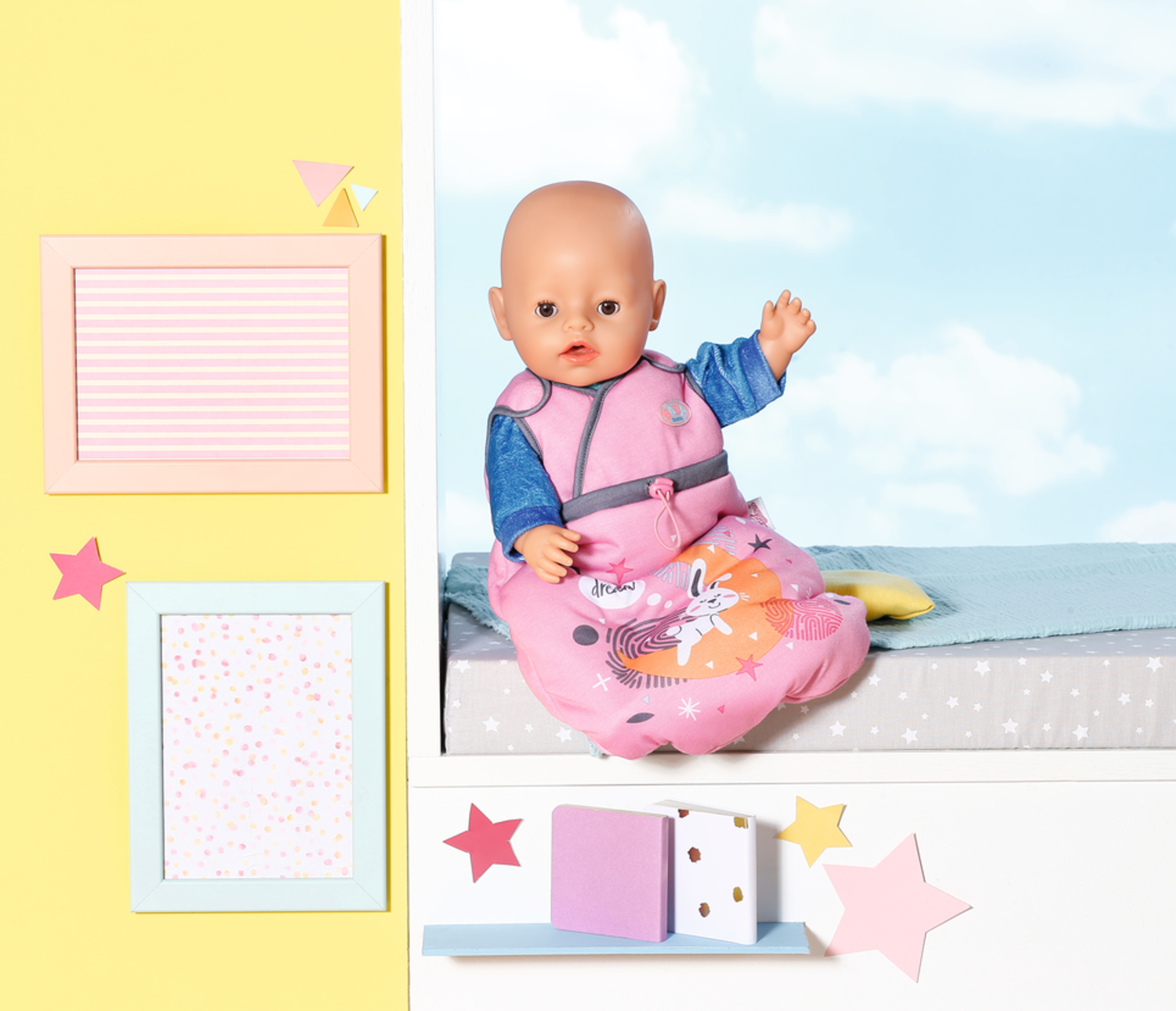 Una muñeca bebé con un saco de dormir rosa está sentada en una superficie. Marcos decorativos y estrellas alrededor, con fondo de cielo azul.