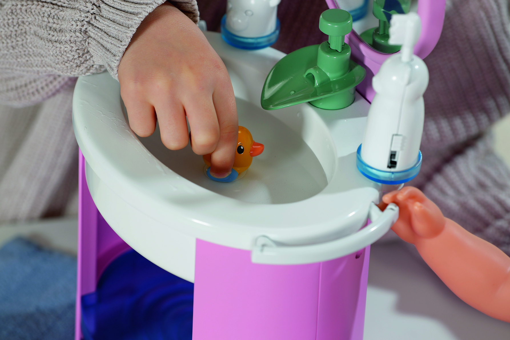 Ręka dziecka umieszcza zabawkę-kaczkę w zabawkowym zlewozmywaku. Zlew jest częścią różowo-białego zestawu zabawek.