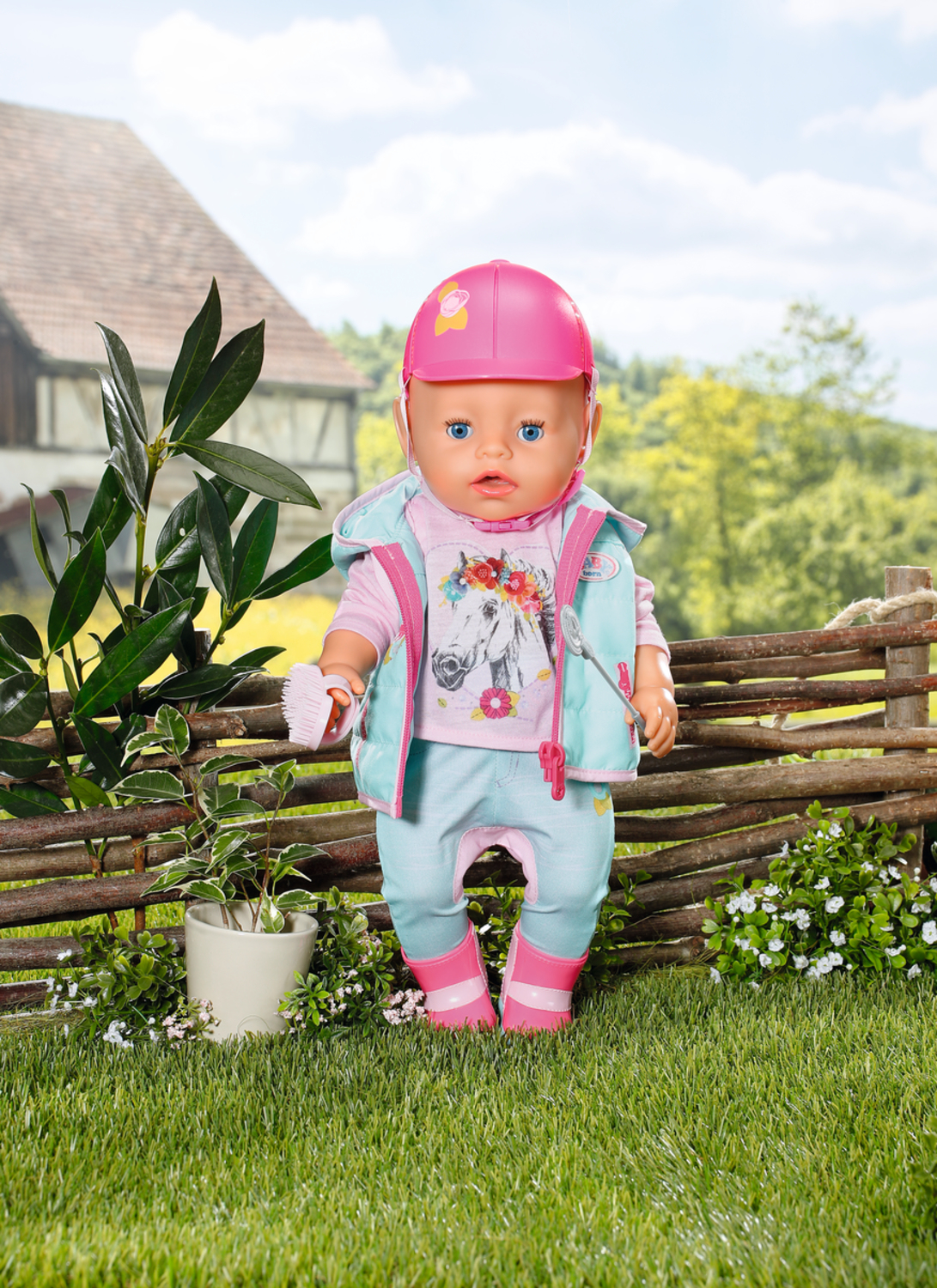 Lalka w stroju jeździeckim z różowym kaskiem, niebieskimi oczami, na trawie. Tło z drewnianym płotem i domem.