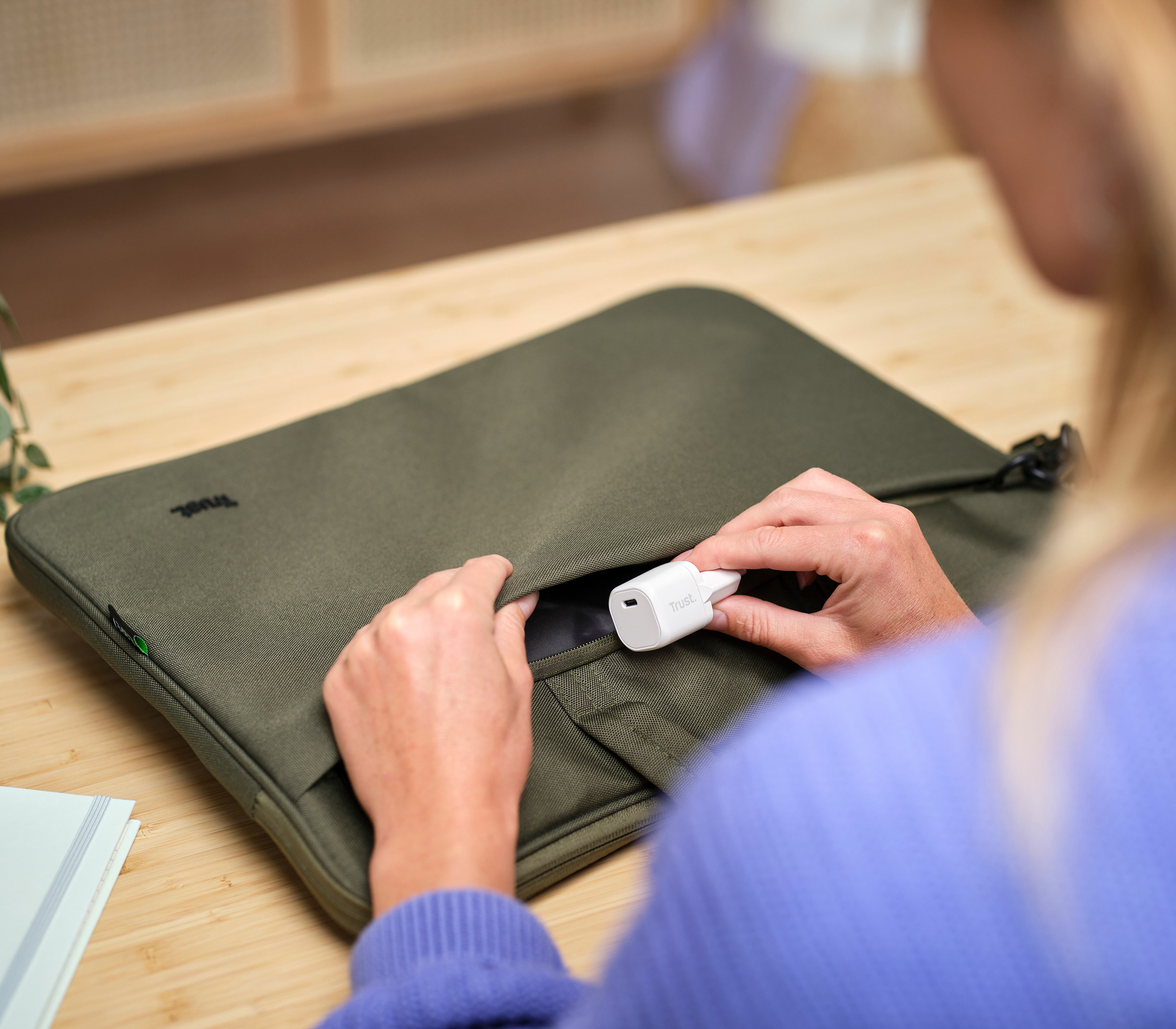 Eine Person steckt ein weißes Ladegerät in eine grüne Laptoptasche. Die Tasche und das Ladegerät haben das Trust-Logo.