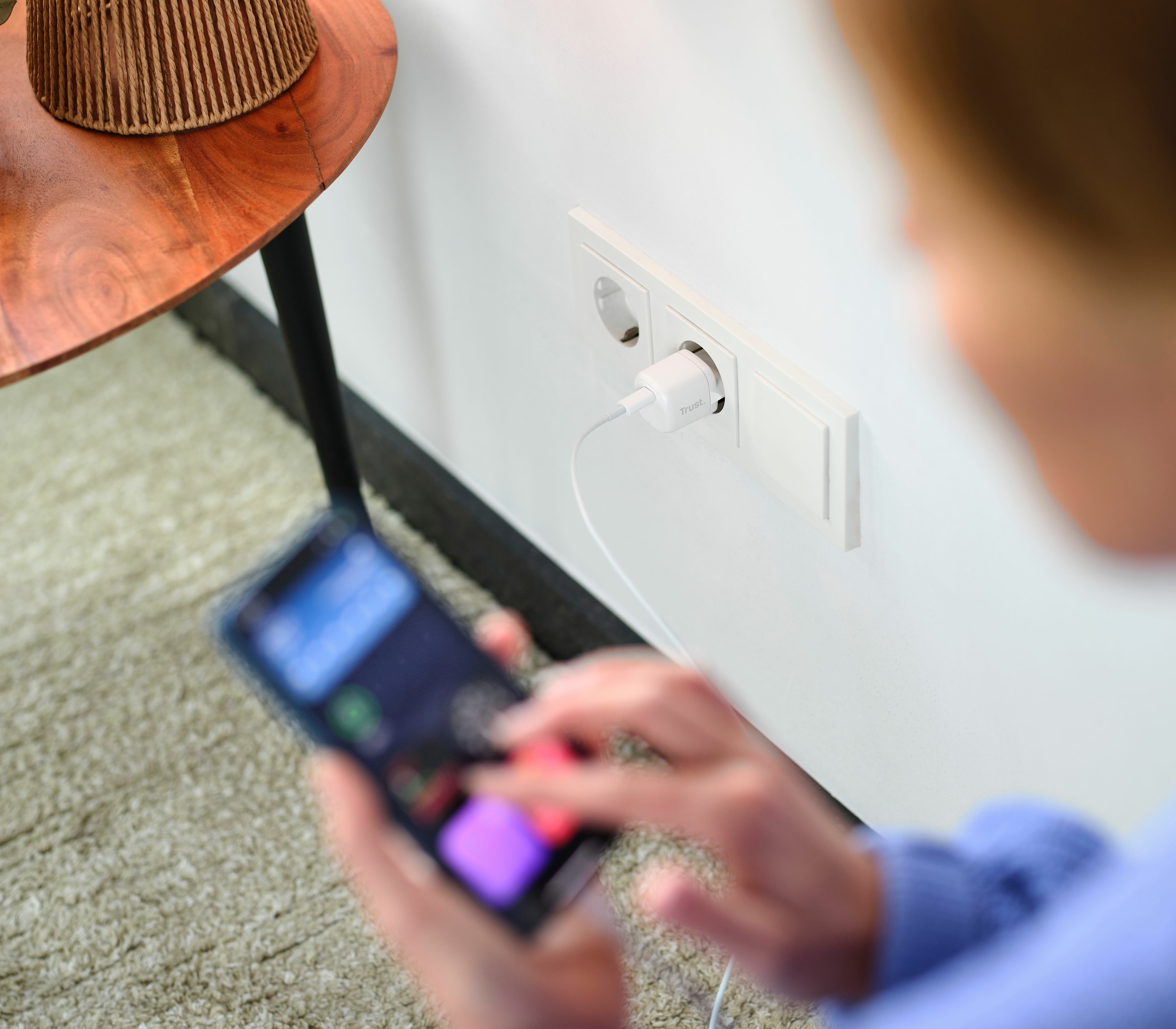 Eine Person benutzt ein Telefon. Ein Ladegerät ist an eine Steckdose angeschlossen. Ein Tisch mit einer Lampe ist im Bild.