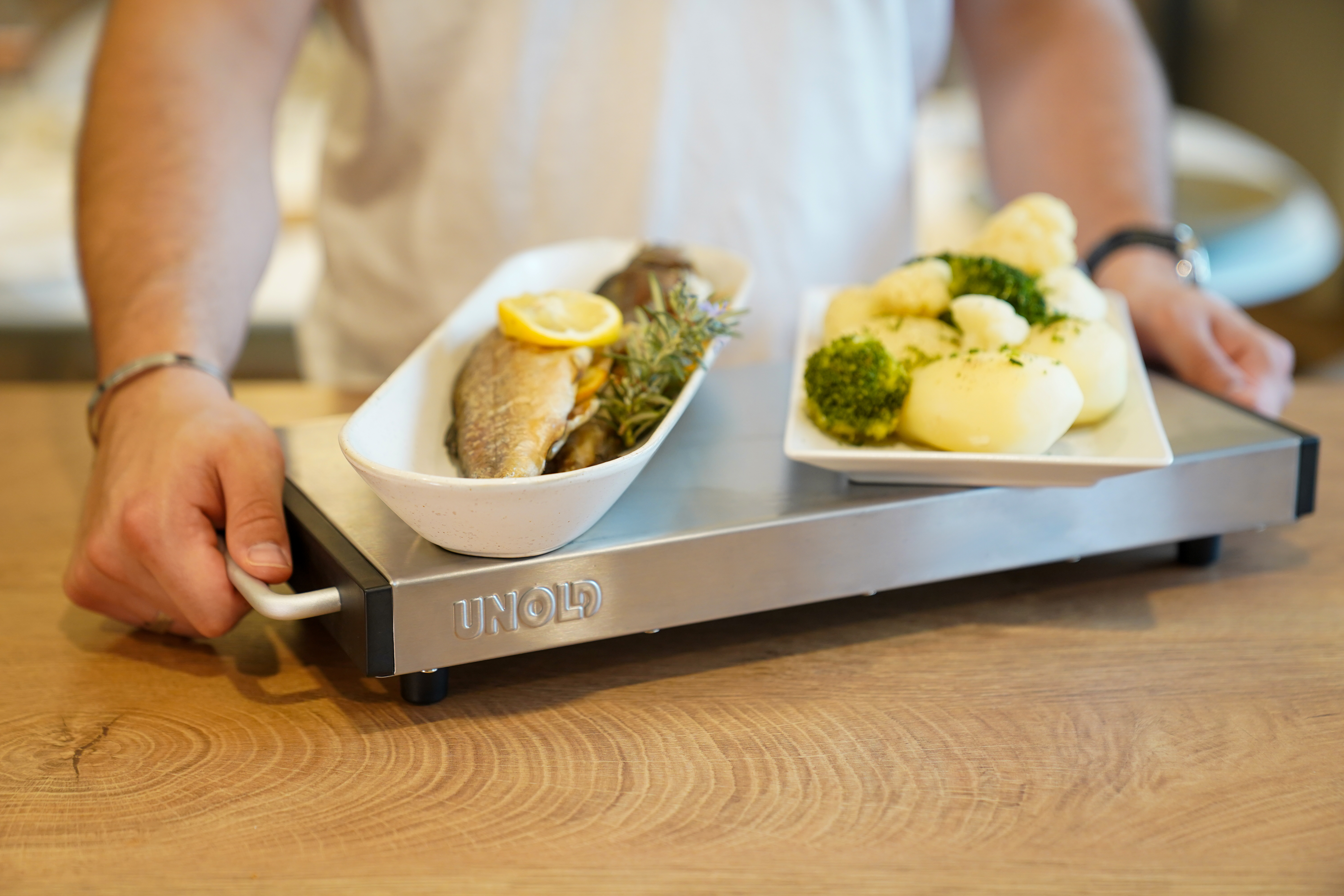 Eine Person hält ein Tablett mit Fisch, Zitrone, Kräutern, Kartoffeln und Brokkoli auf einem Holztisch.