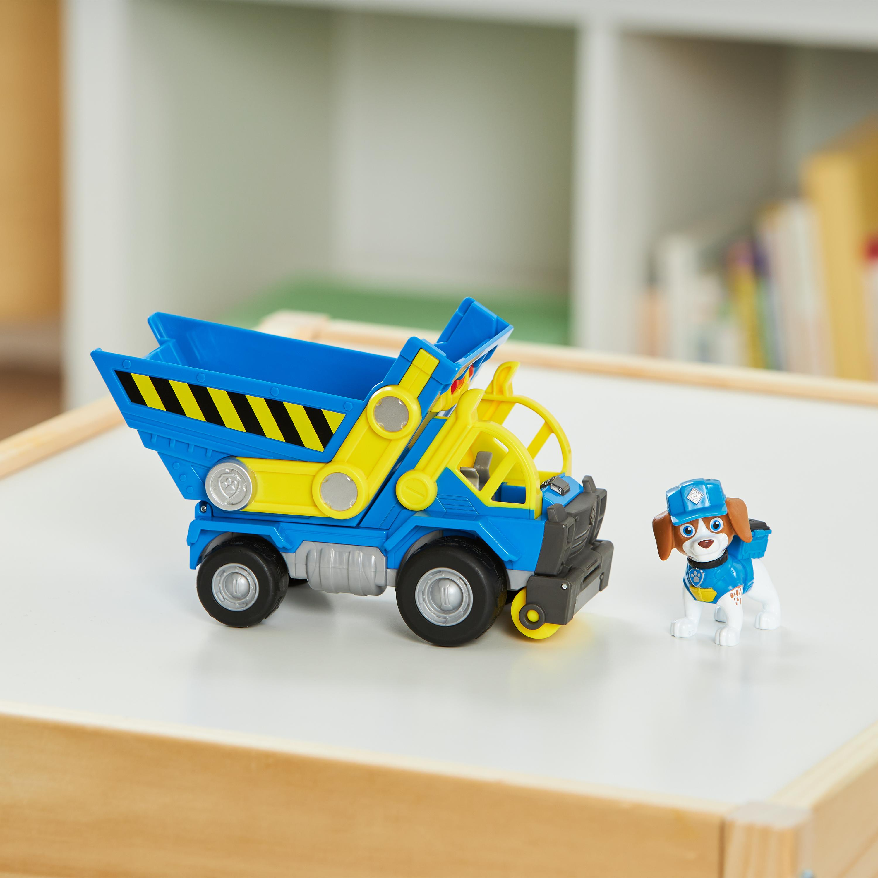 Ein blau-gelber Spielzeug-Muldenkipper und eine Hundefigur auf einer weißen Oberfläche. Hintergrund: Regal und Bücher.
