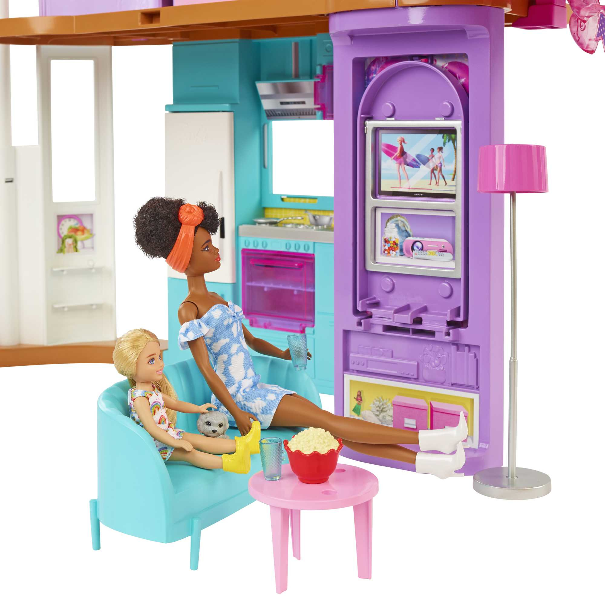 Ein Barbie-Spielhaus mit Puppen und Zubehör. Puppen sitzen auf einem blauen Sofa, ein Tisch hat Popcorn. TV zeigt Surfer.