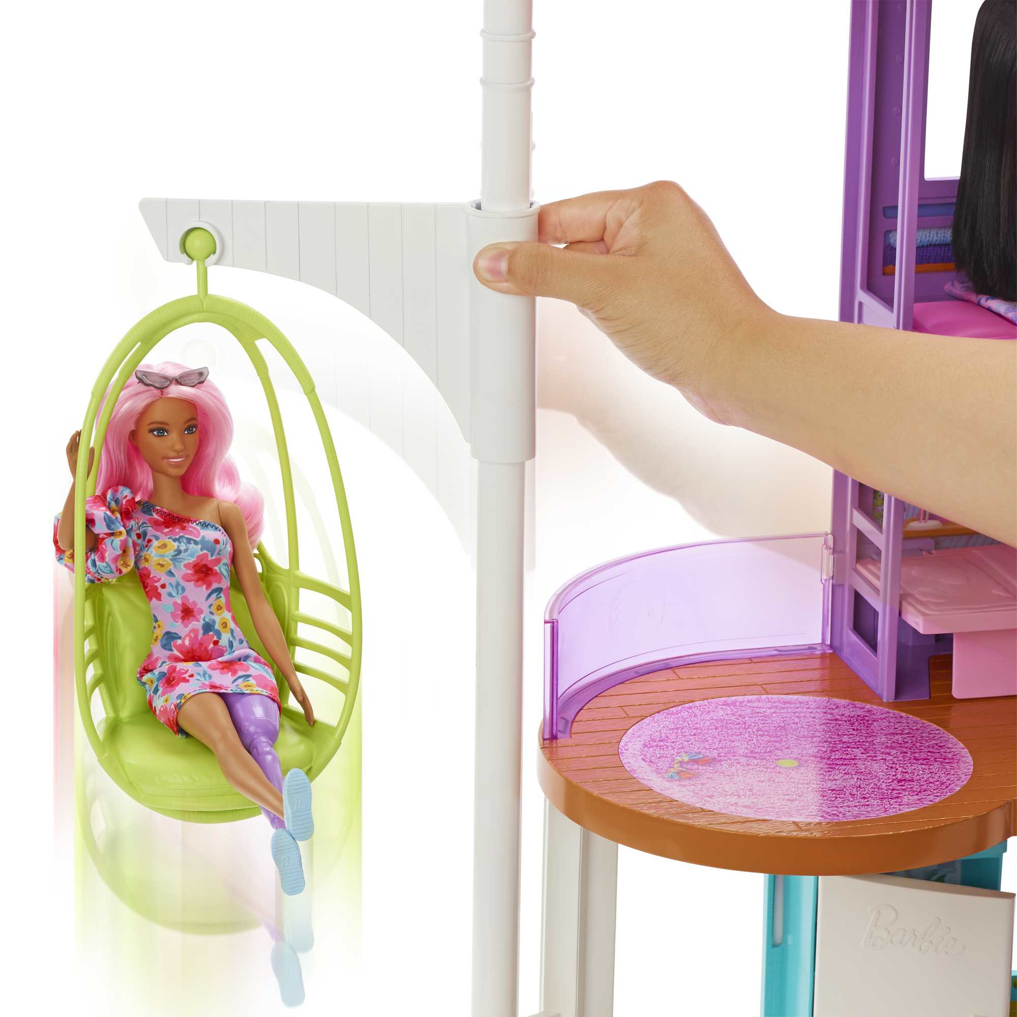 Eine Barbie-Puppe mit rosa Haaren sitzt in einer grünen Schaukel. Eine Hand justiert die Haltestruktur. Ein violetter Spielbereich ist sichtbar.