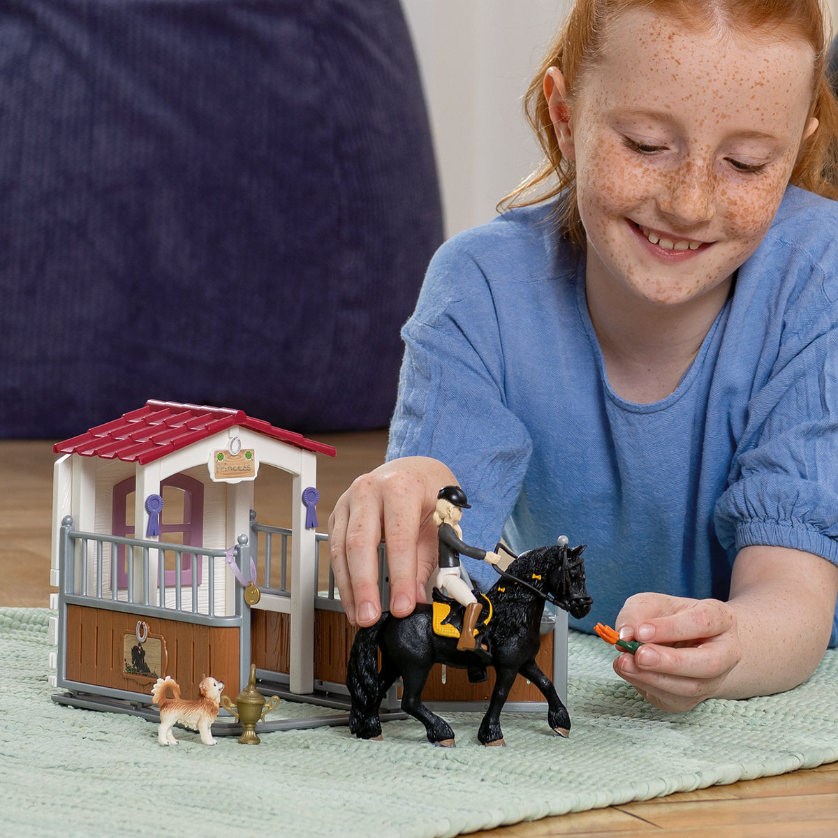 Ein Mädchen spielt mit Pferdespielzeug. Ein schwarzes Pferd mit Reiter, ein Stall, ein Hund und eine Trophäe auf einer grünen Matte.