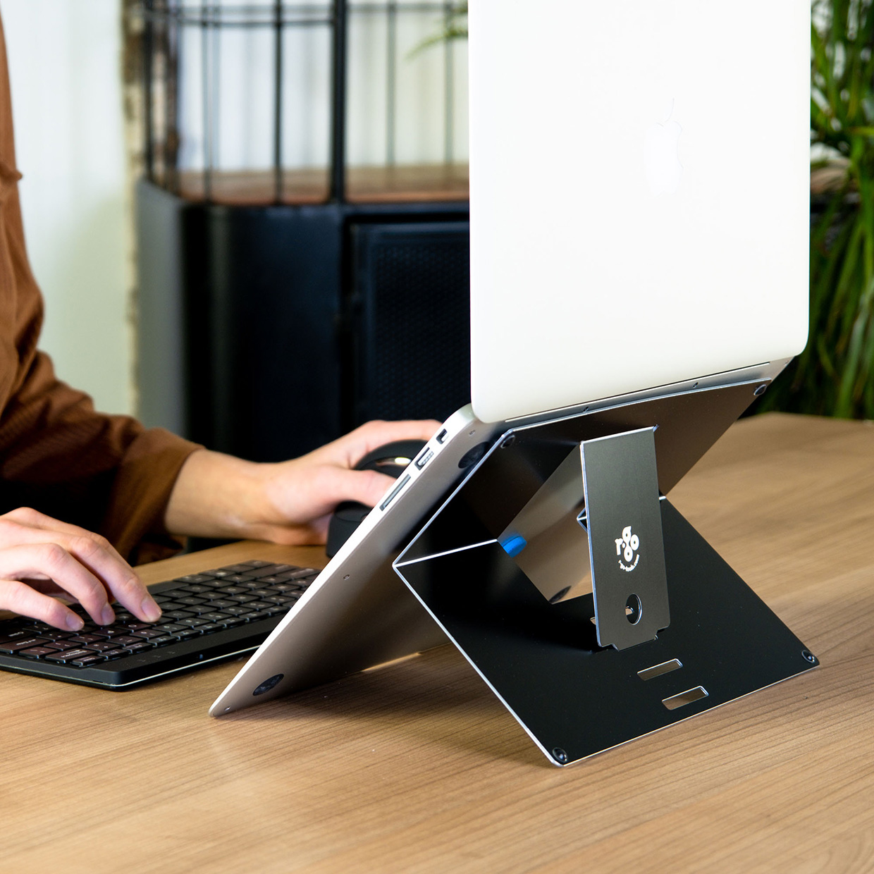 Ein Laptop steht auf einem schwarzen Ständer. Eine Person benutzt Tastatur und Maus. Im Hintergrund sind Tisch und Schrank.