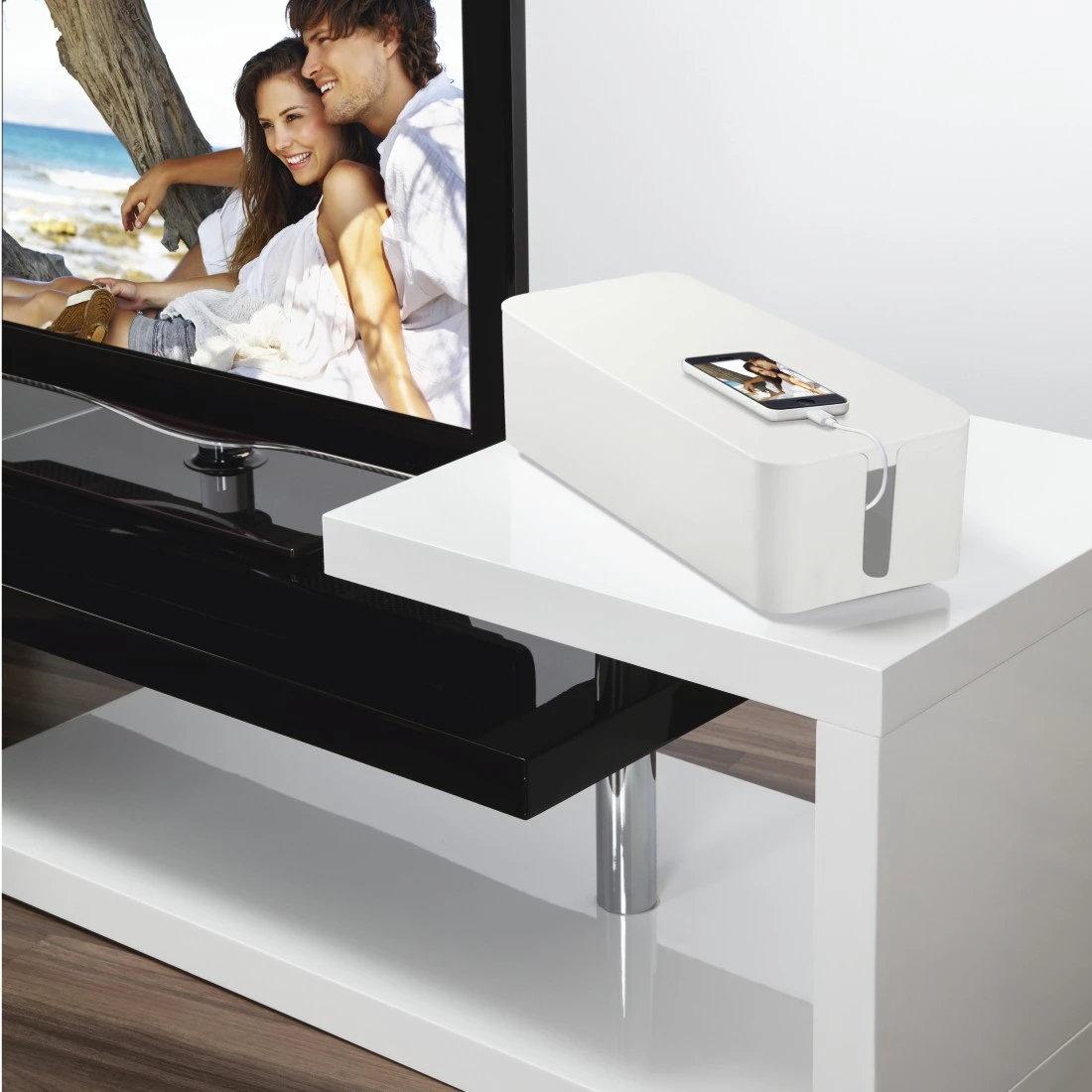 Una scatola per cavi bianca su un tavolo bianco. Un telefono è appoggiato sulla scatola, con il cavo di ricarica collegato. Una TV mostra una coppia.