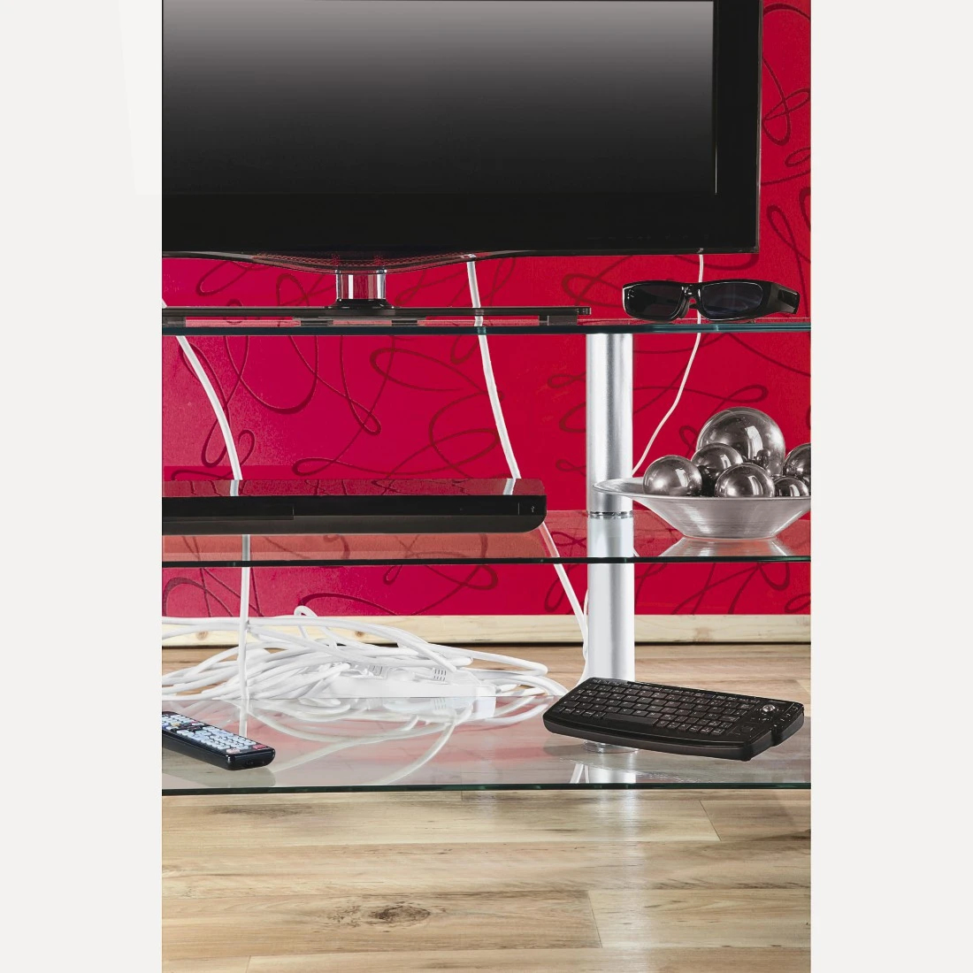 TV su un supporto in vetro con accessori. Parete rossa con vortici sullo sfondo. Tastiera, telecomando e cavi sul supporto.