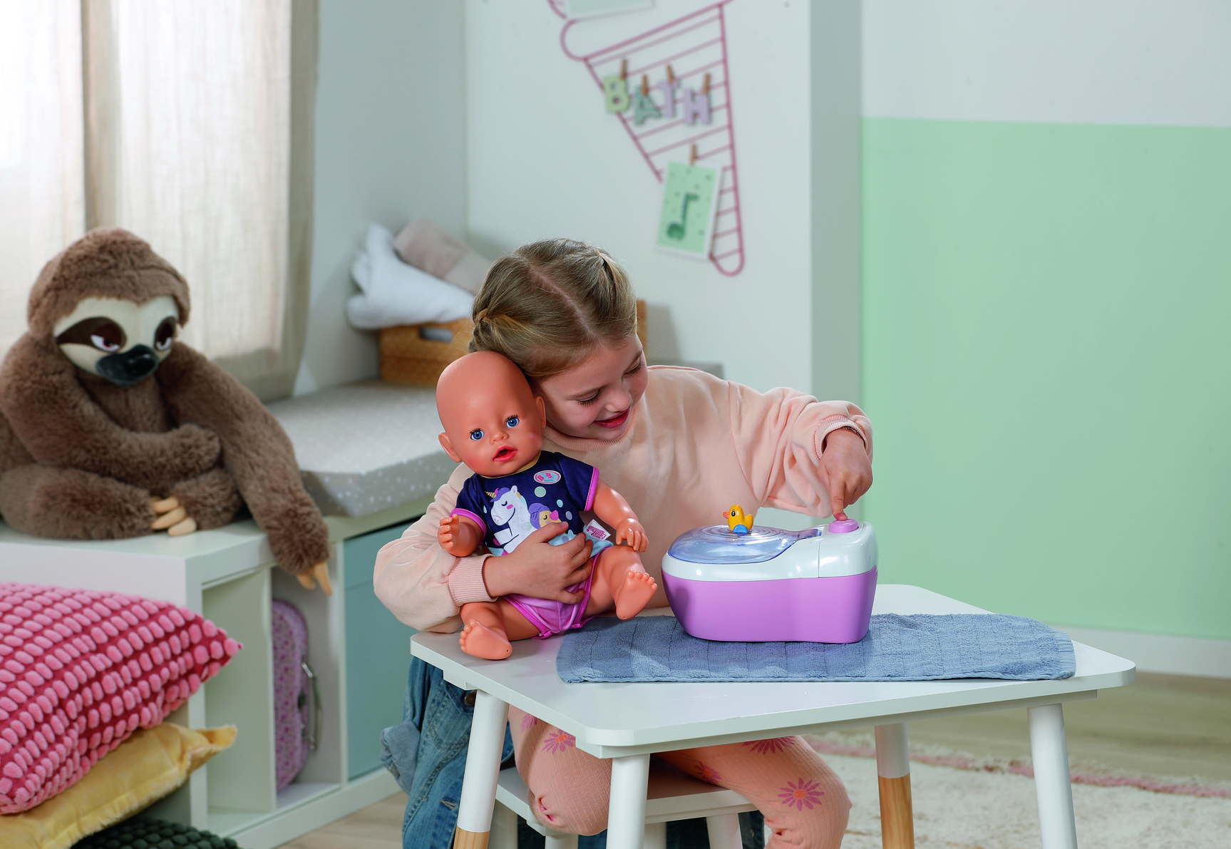 Dziewczynka bawi się lalką i zabawką. Pluszowy leniwiec siedzi na półce. Różowo-biała zabawka stoi na białym stole.
