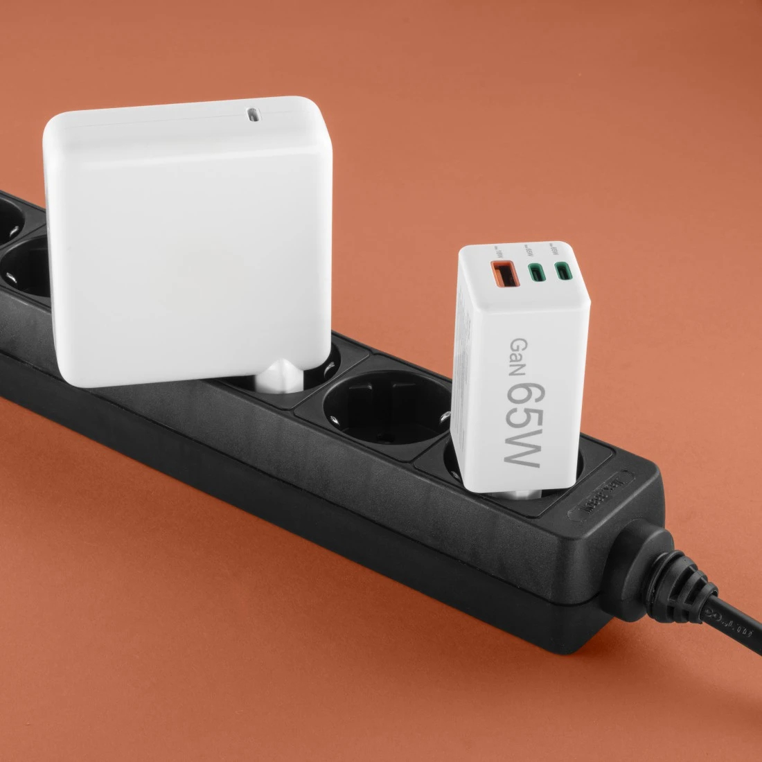 Zwei weiße Netzadapter, die in eine schwarze Steckdosenleiste auf orangefarbenem Hintergrund eingesteckt sind. Ein Adapter hat 'Gan 65W' drauf.