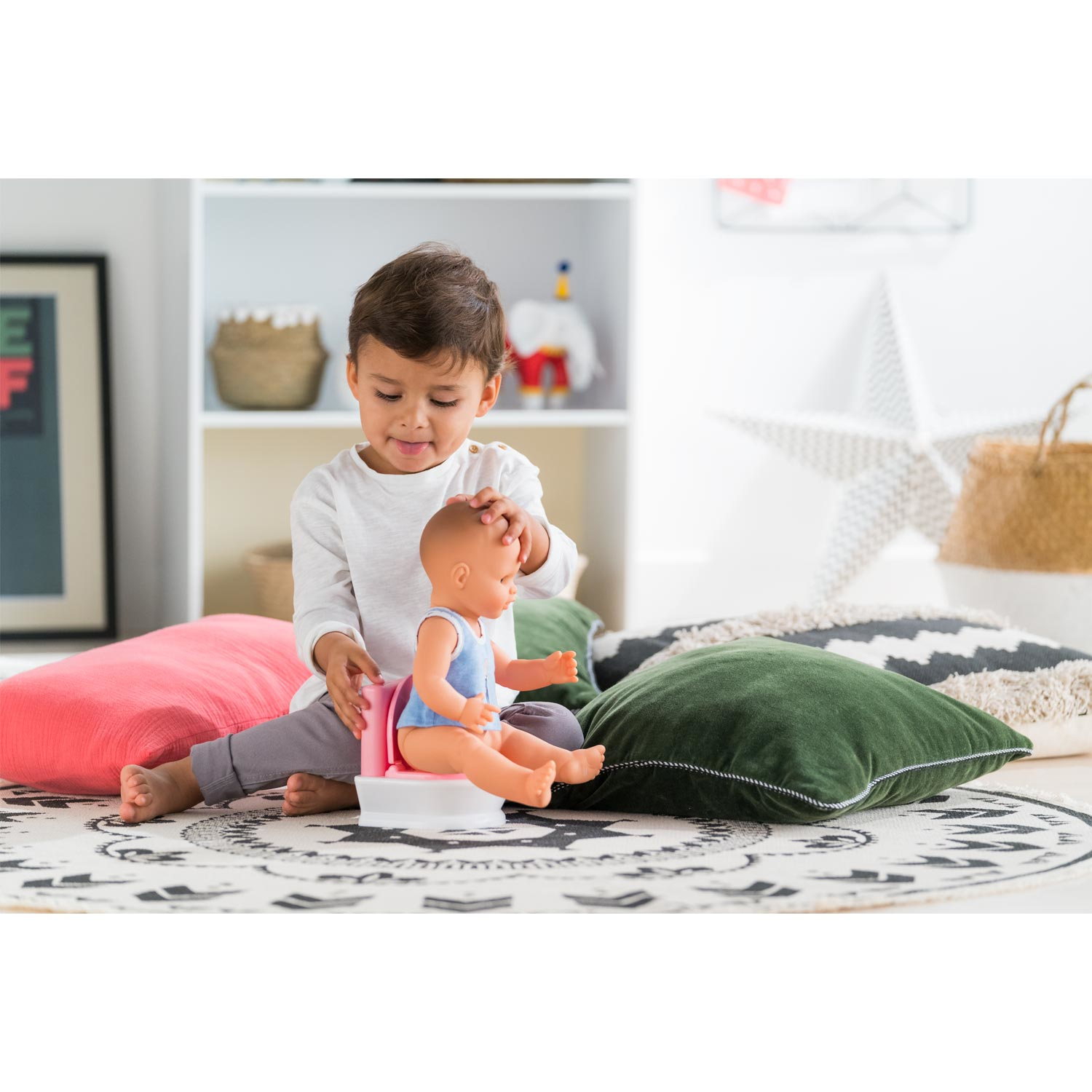 Dziecko bawi się lalką na nocniku. Siedzą na wzorzystym dywanie otoczeni poduszkami. Półka i inne przedmioty w tle.