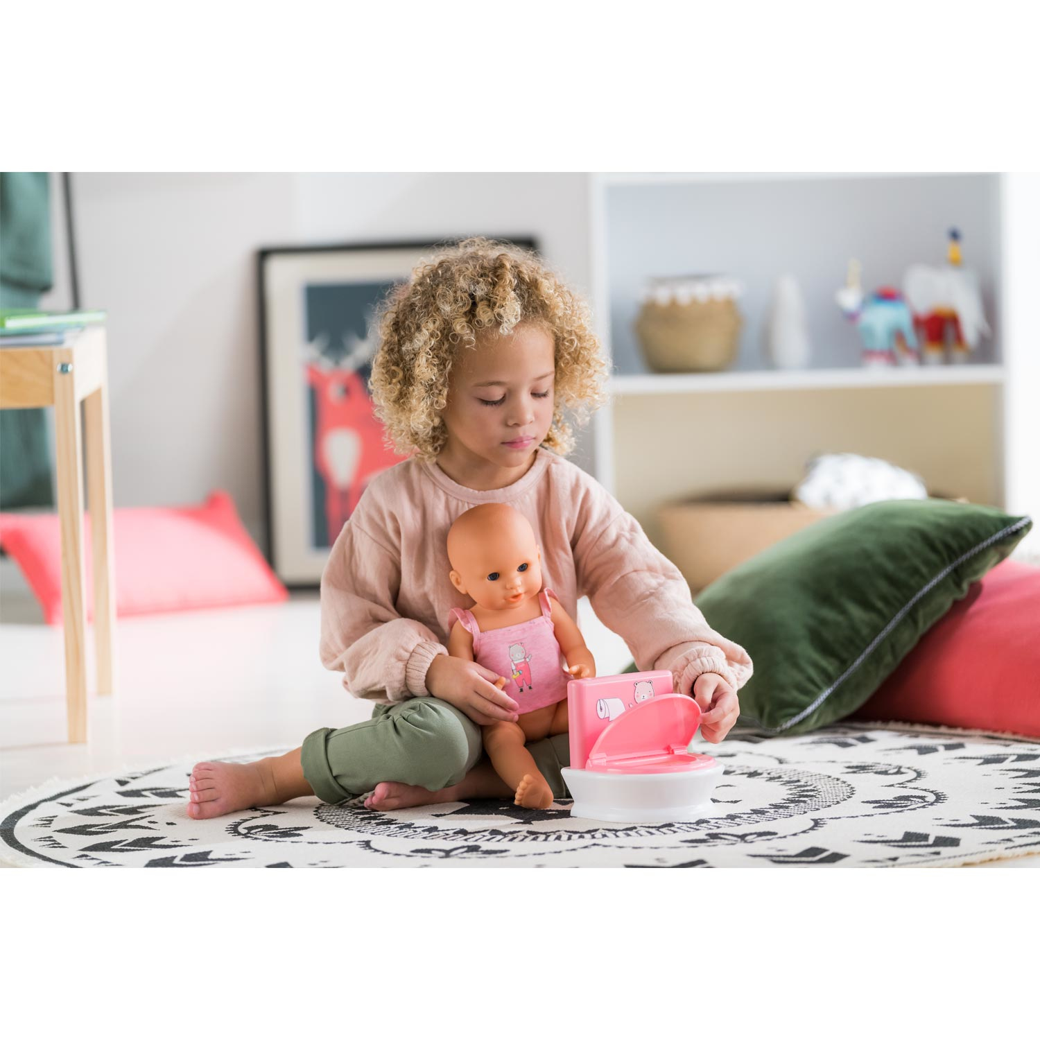 Dziecko z kręconymi włosami siedzi trzymając lalkę. Dziecko bawi się toaletą-zabawką. Pod spodem jest dywan z czarno-białym wzorem.