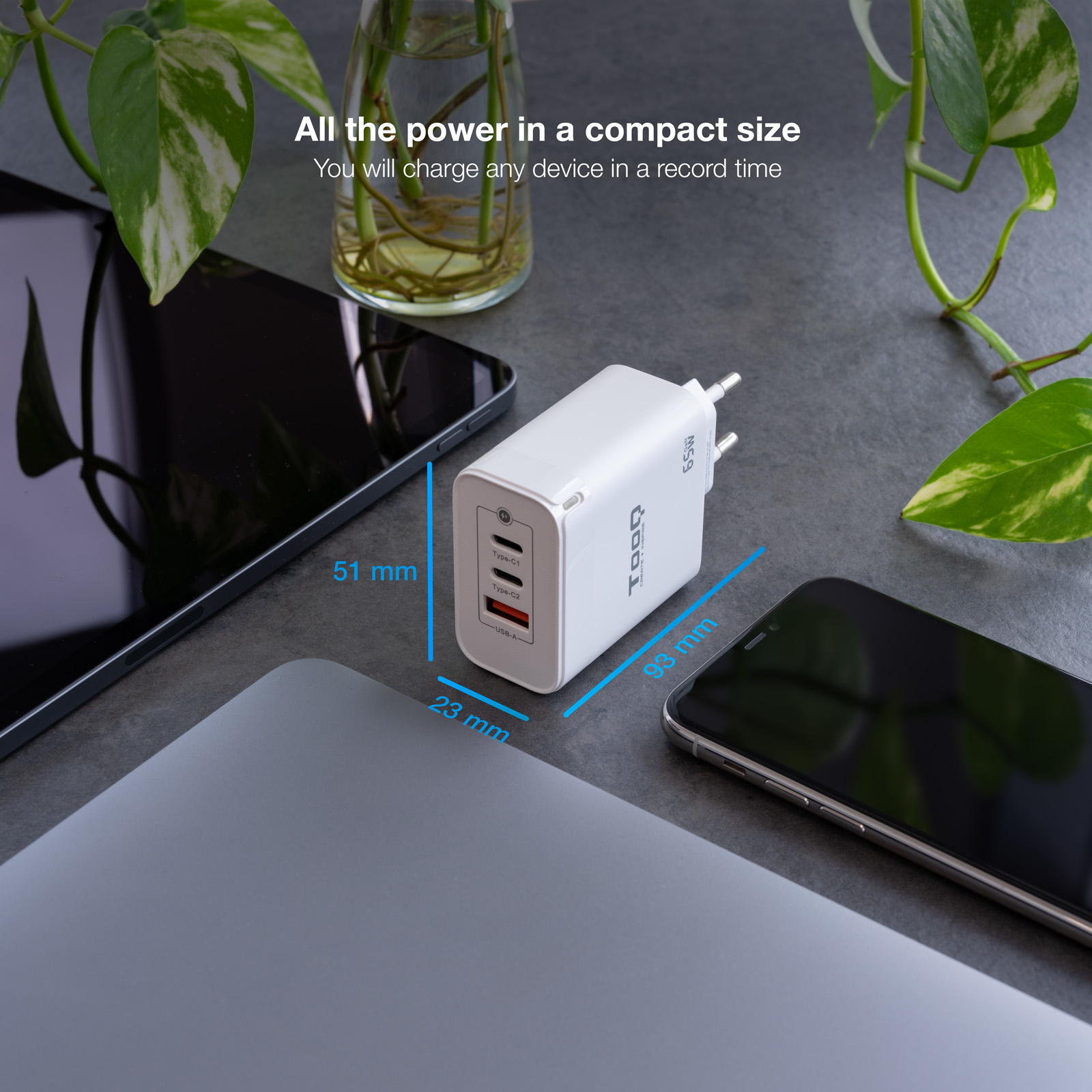 Un cargador blanco con dimensiones, cerca de una tableta, teléfono y portátil. Plantas verdes en el fondo.