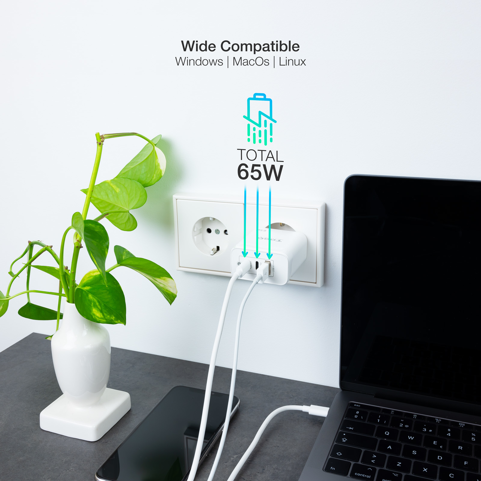 Un adaptador de corriente blanco enchufado a una toma de corriente, cargando una computadora portátil y un teléfono. Una planta en un jarrón está a la izquierda.