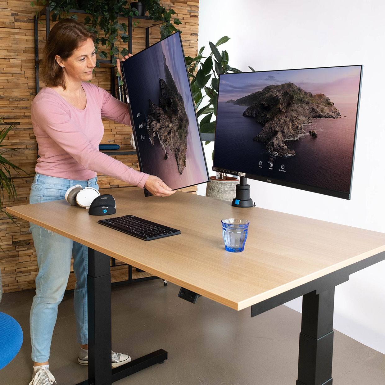 Woman setting up a monitor on a desk. Another monitor is on the right. Keyboard, mouse, and glass are on the desk. Brick wall in the back.