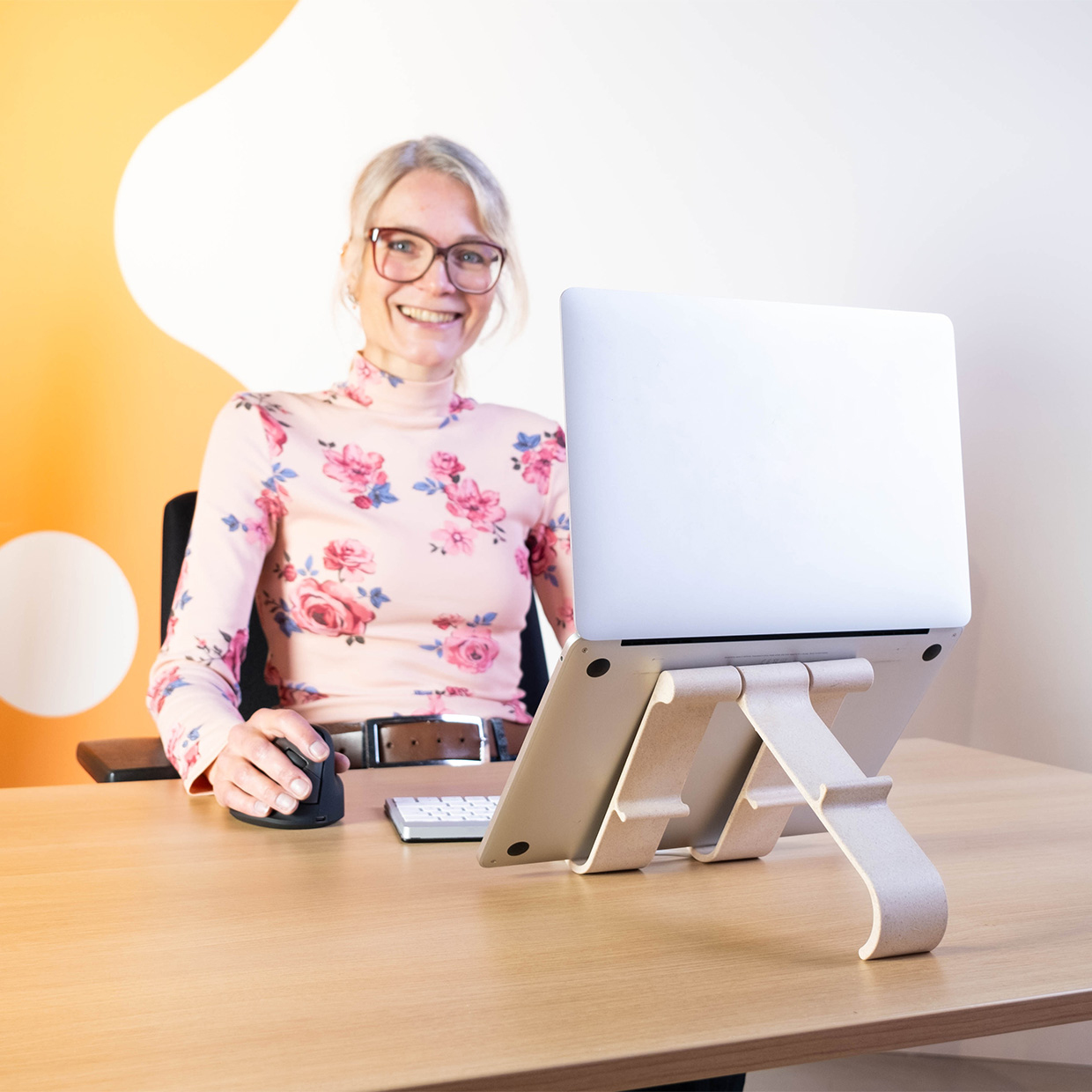 Woman with glasses smiles while working on a laptop. The laptop is on a stand on a wooden desk. Flowers on her shirt.