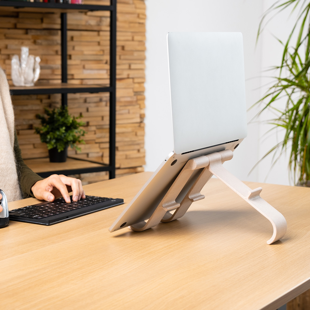 A laptop on a stand sits on a wooden desk. A person uses a keyboard. A bookshelf and plant are in the background.