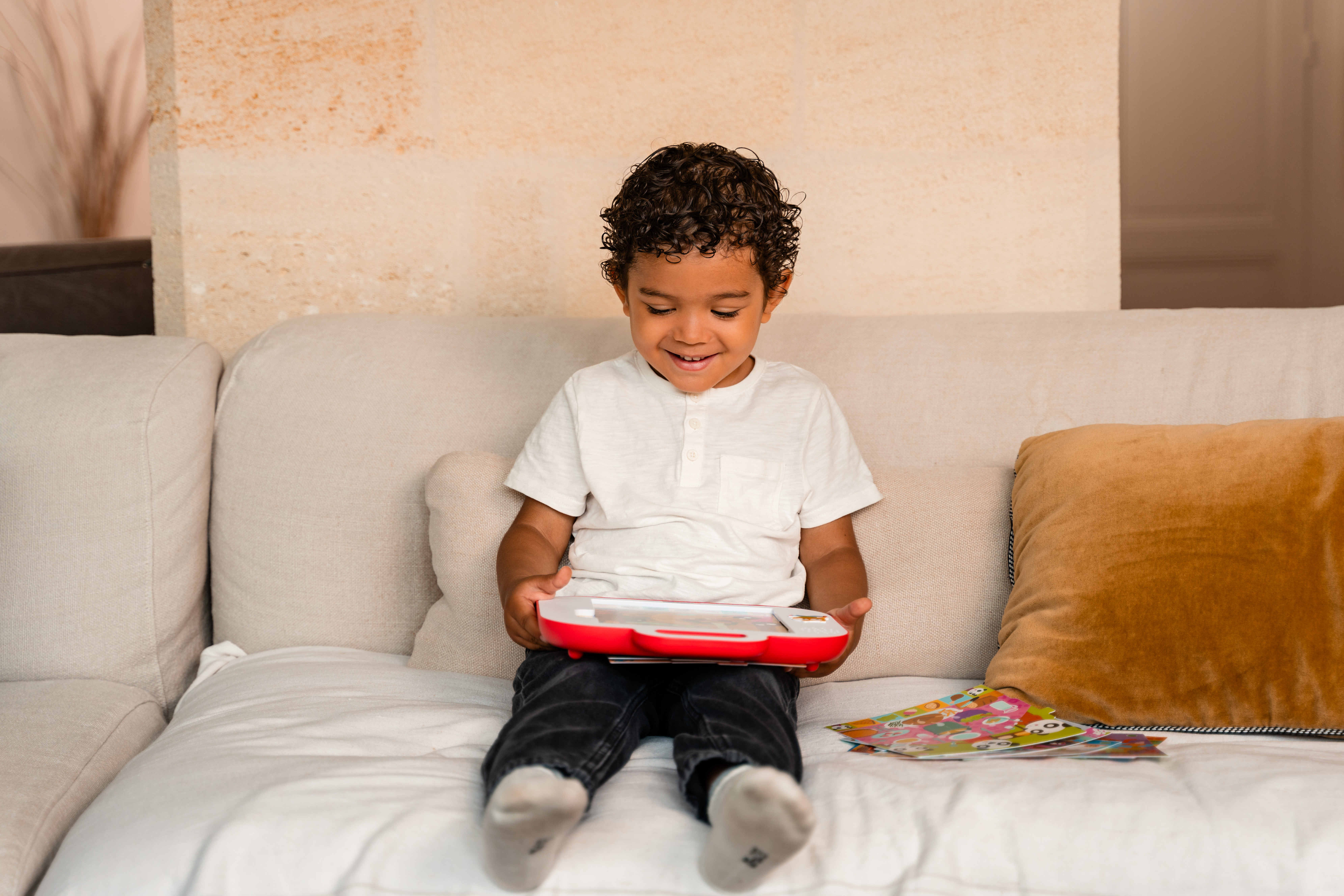 Ein lächelnder Junge sitzt auf einem Sofa und hält ein rotes Spielzeug. Bunte Bilder liegen neben ihm. Rechts ist ein Kissen.