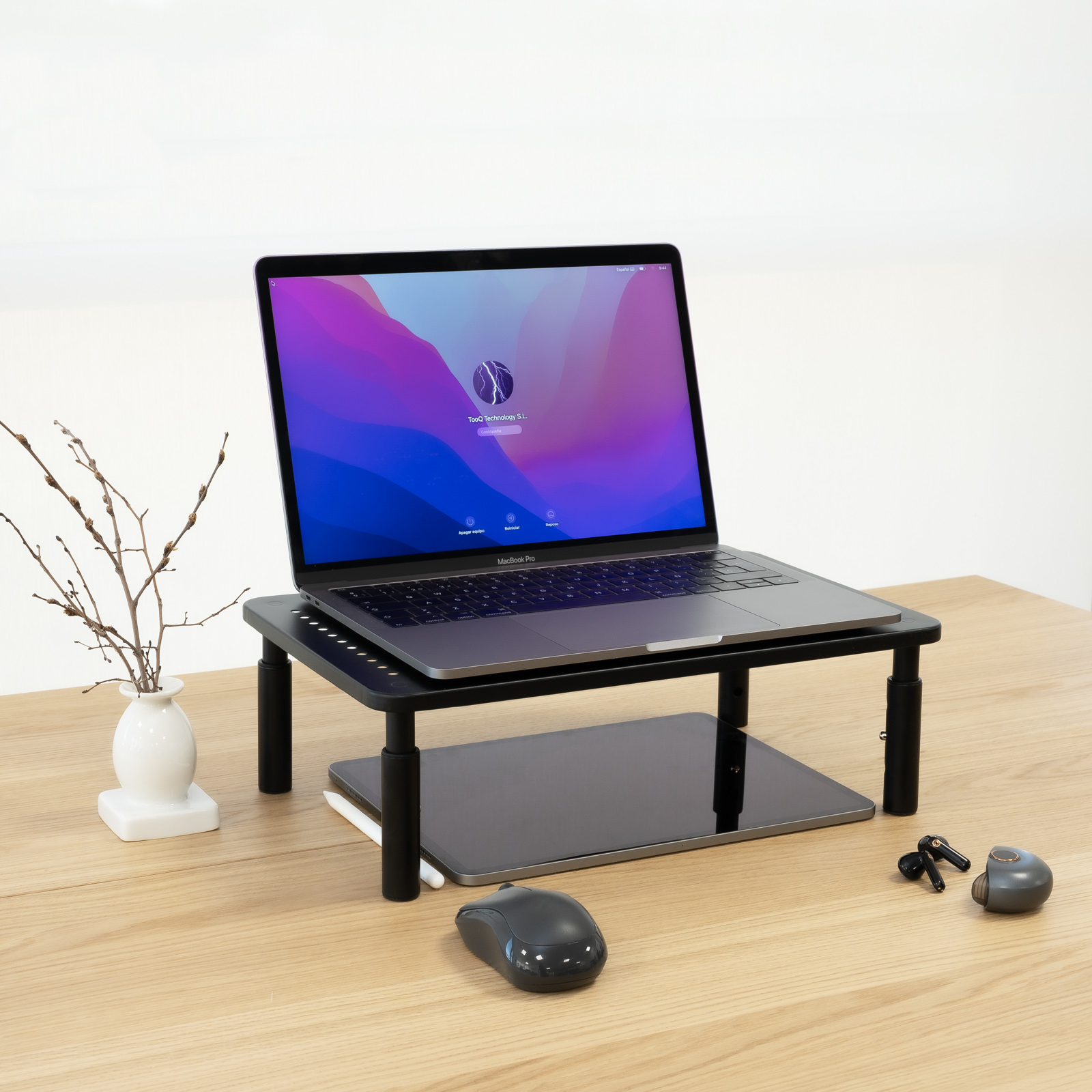 Laptop su supporto, tablet, mouse e auricolari su scrivania in legno. Un piccolo vaso con rami a sinistra.