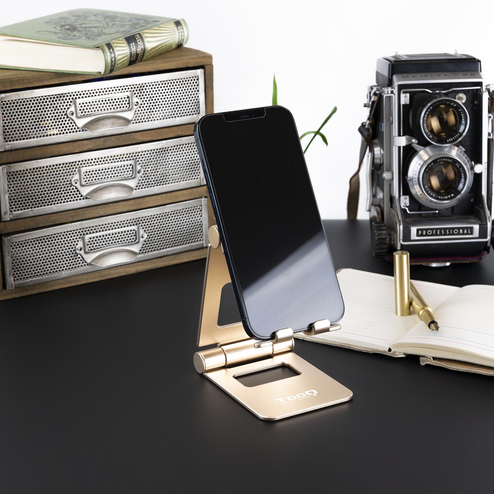Soporte dorado para teléfono con teléfono negro, cámara vintage, cuaderno y cajones sobre una superficie negra.