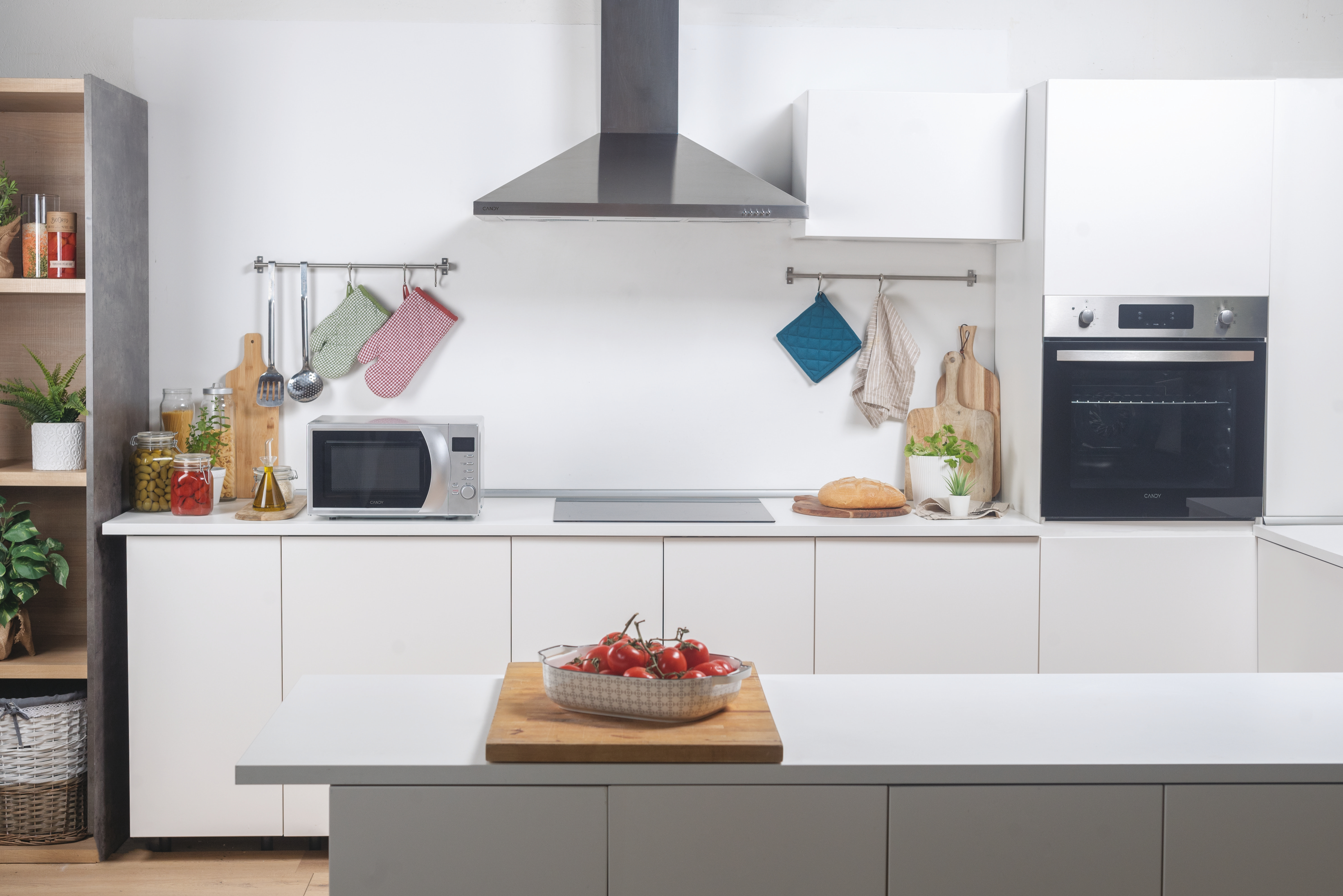 Una cucina moderna con mobili bianchi, elettrodomestici e un tavolo con pomodori. Utensili appesi sopra.